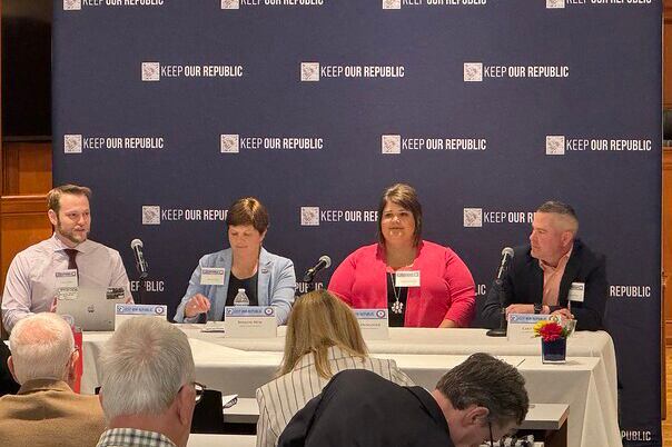 Four people in business clothes sit at a table in front of a large group of people.