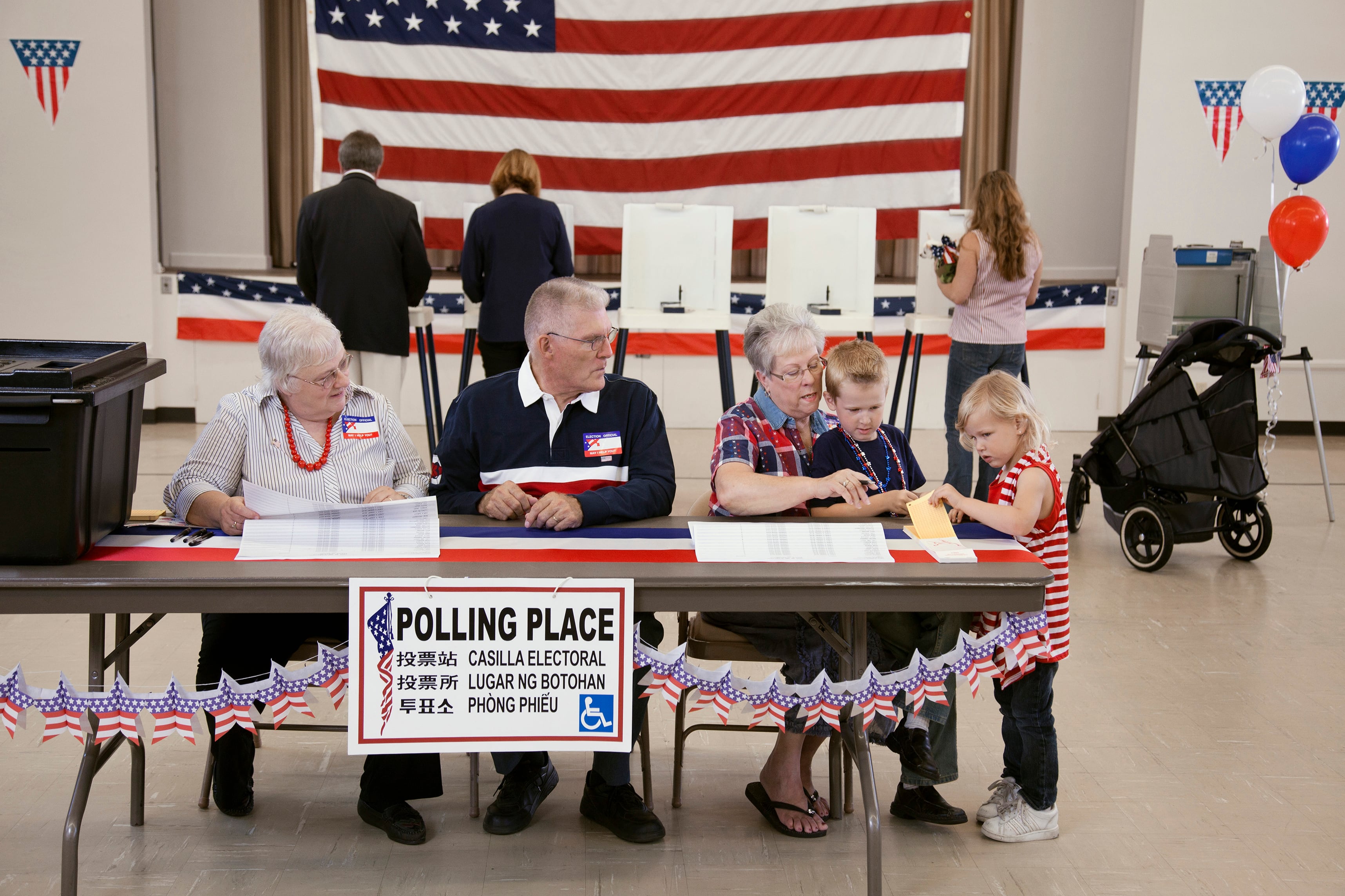 Three older adults sit at a table while two children stand nearby all inside a building at a polling location.