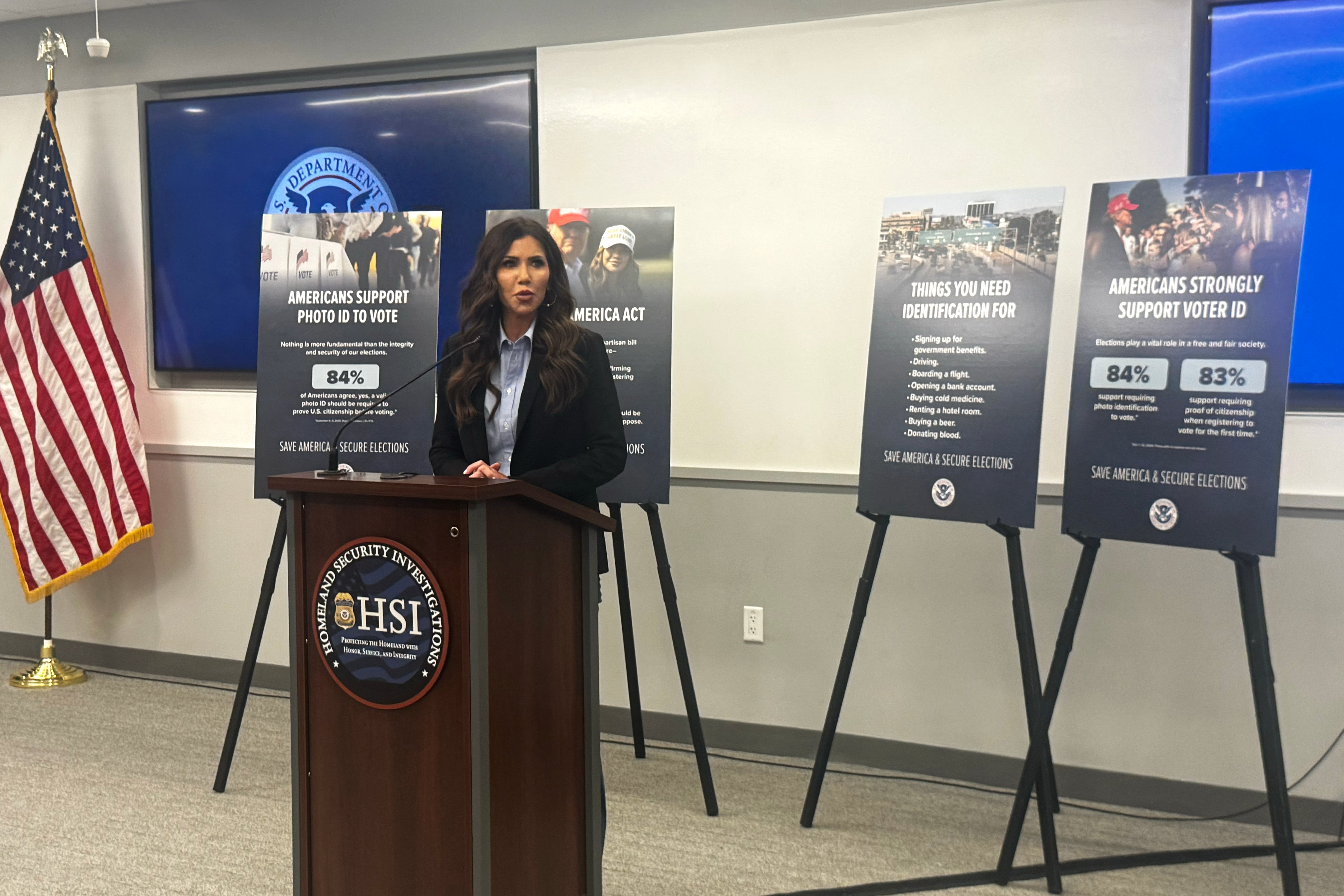 A photograph of a white woman in a suit standing behind a wooden podium and in front of four poster boards on stands in a conference room.