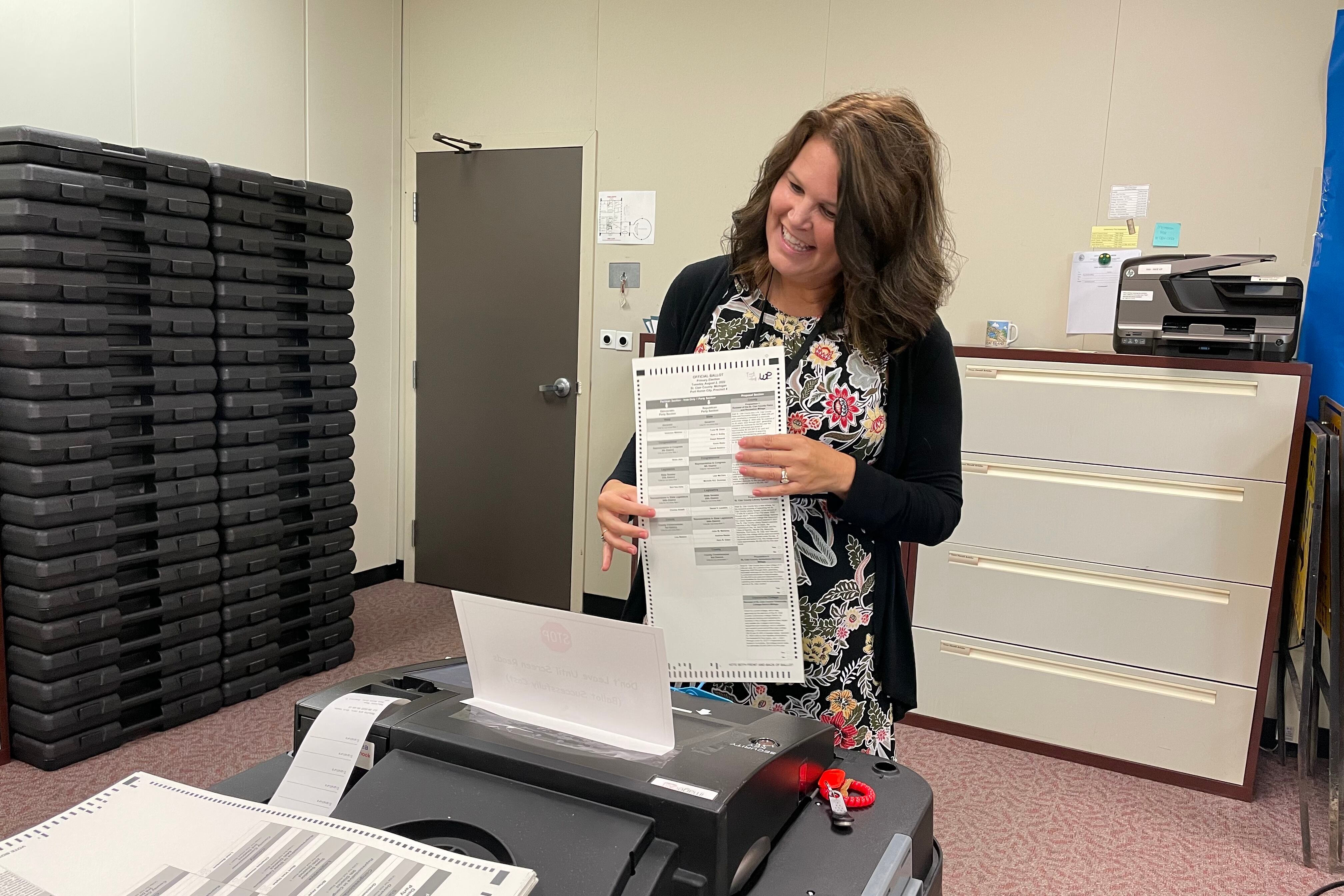 A woman in an office stands before a scanner holding a ballot 