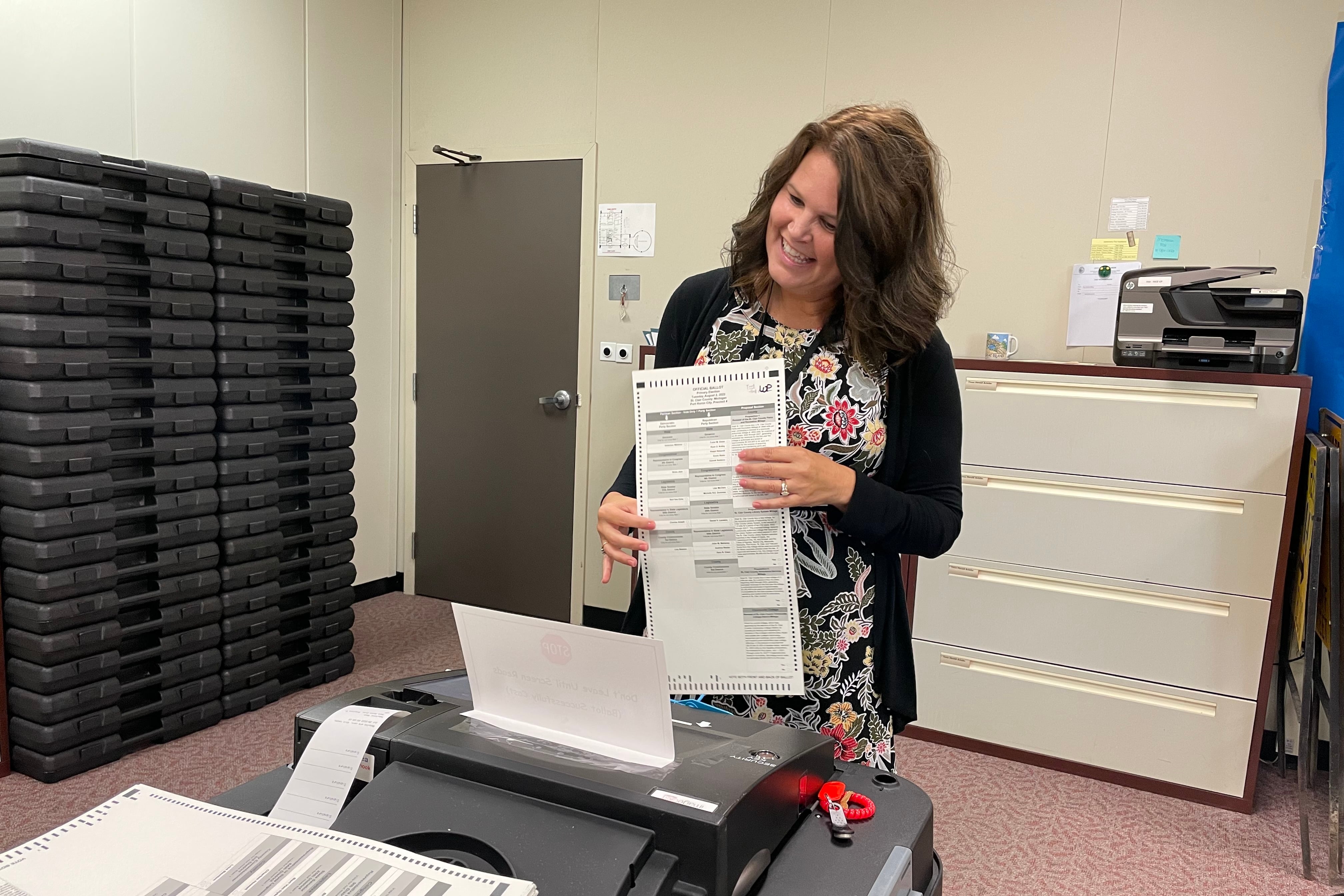 A woman in an office stands before a scanner holding a ballot