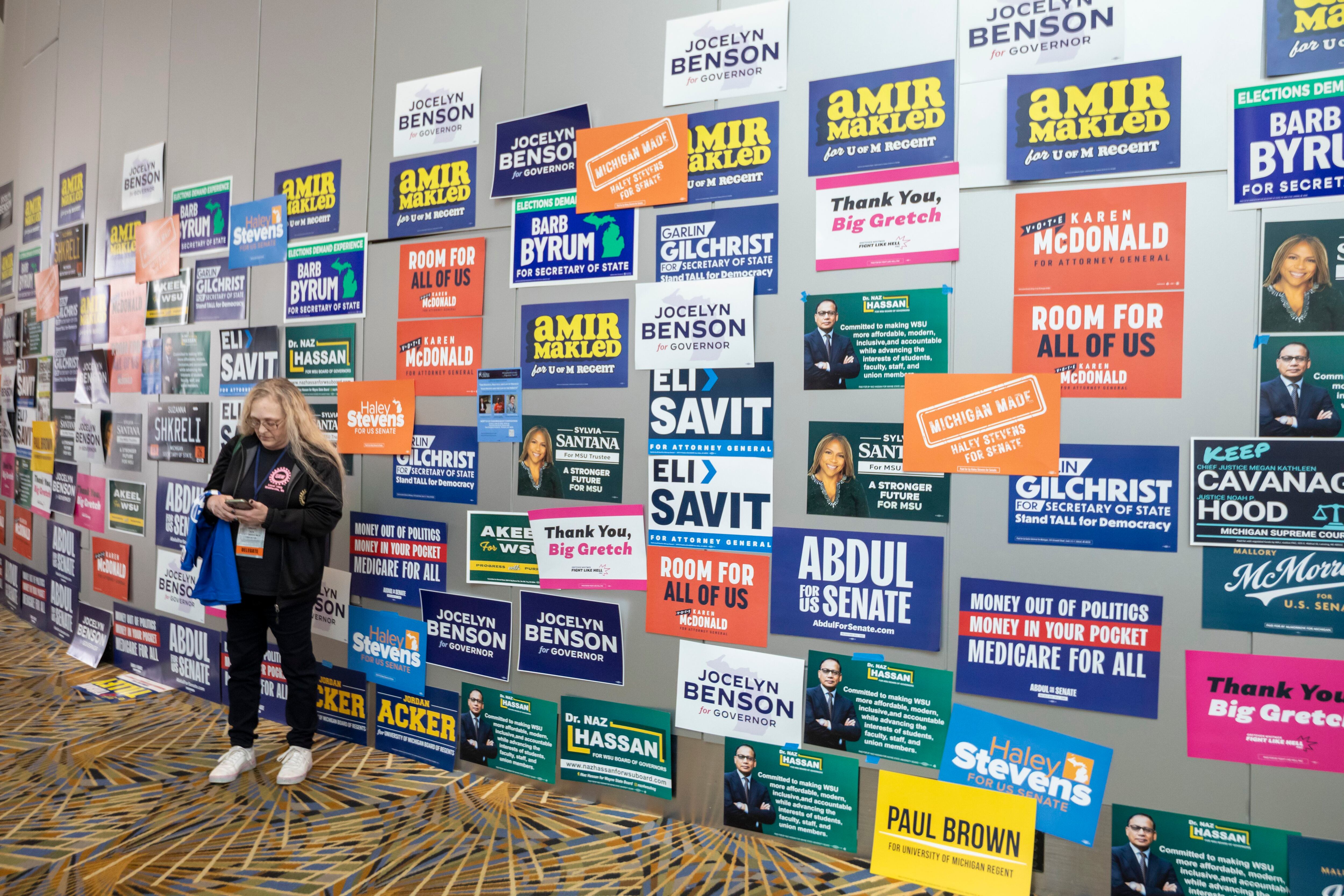 A photograph of a person standing in front of a large convention center wall covered in campaign posters.
