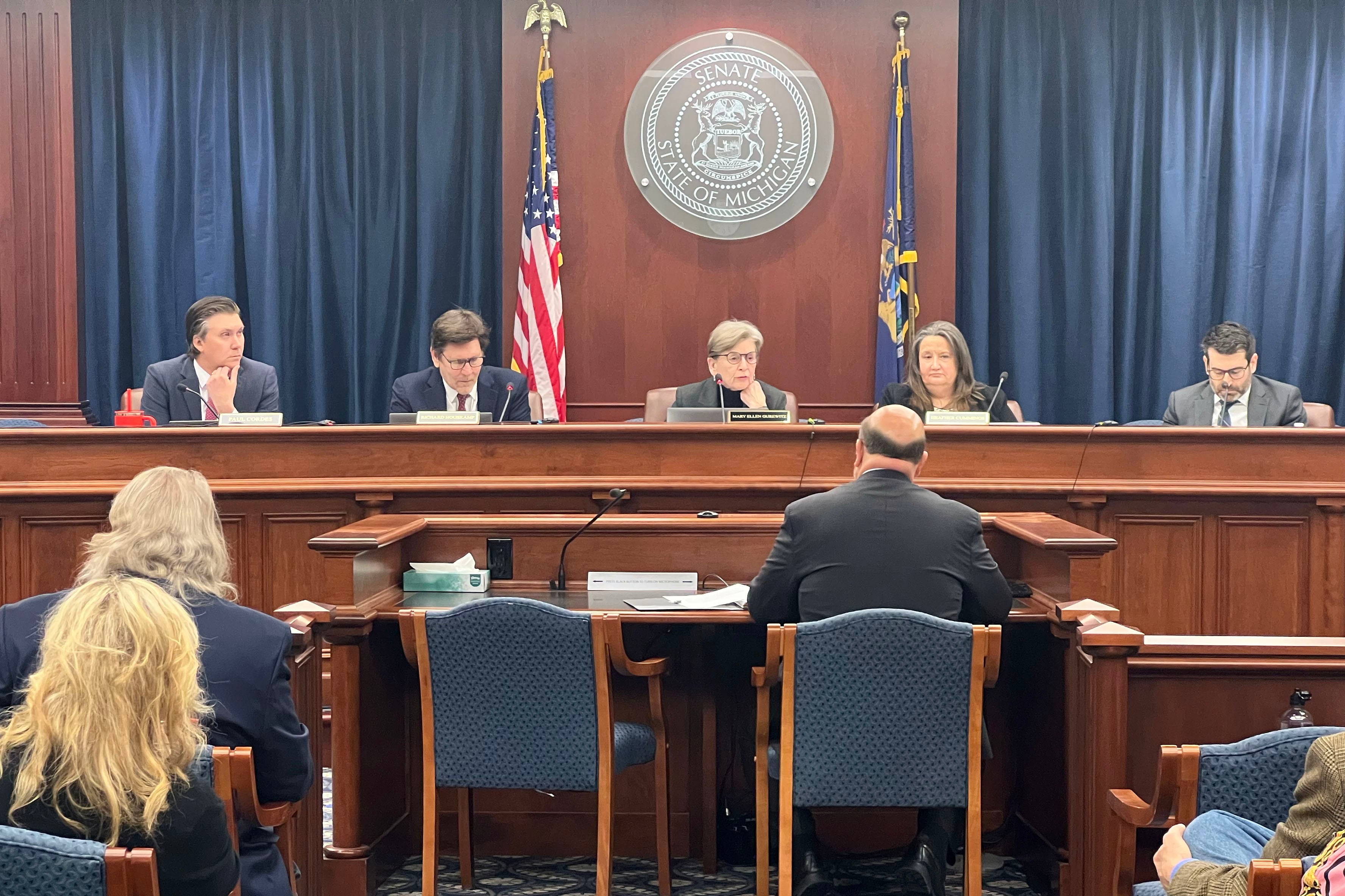 A group of people in suits sit at a long wooden table in a state building conference room.