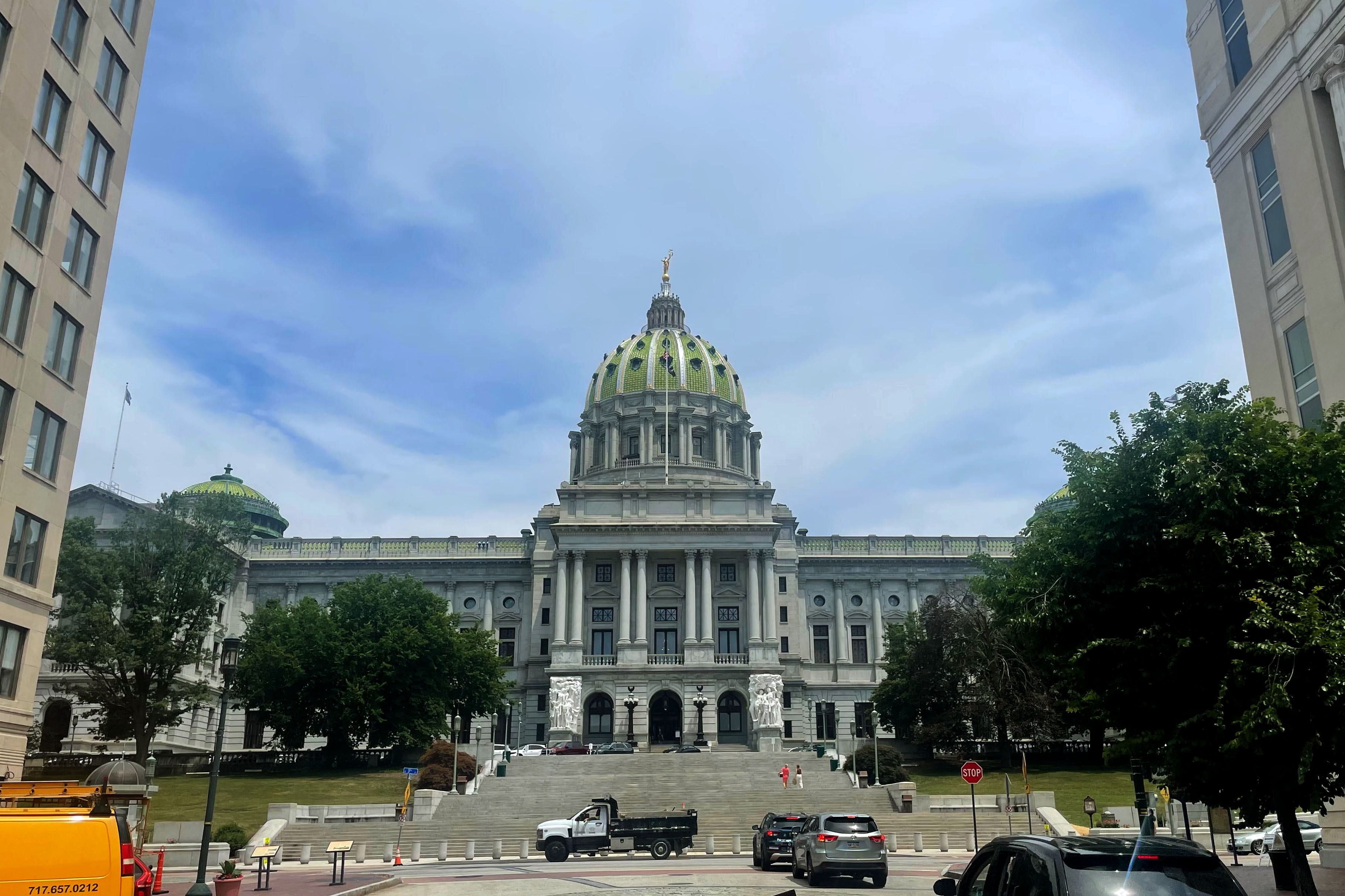 A large white stone state Capitol building with a blue sky with clouds and some cars are out front.