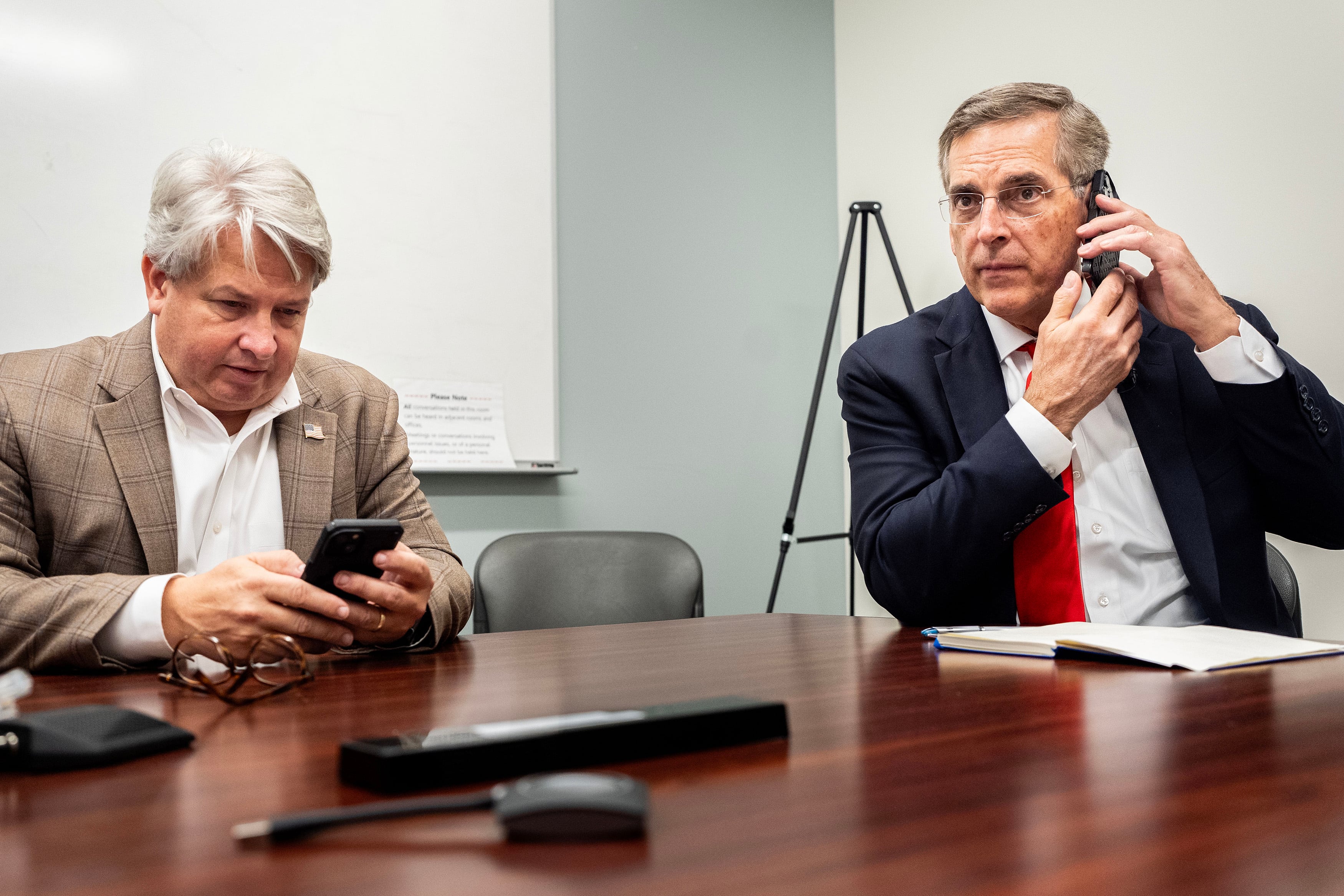 Two men in suits sit at a wooden table in an office.