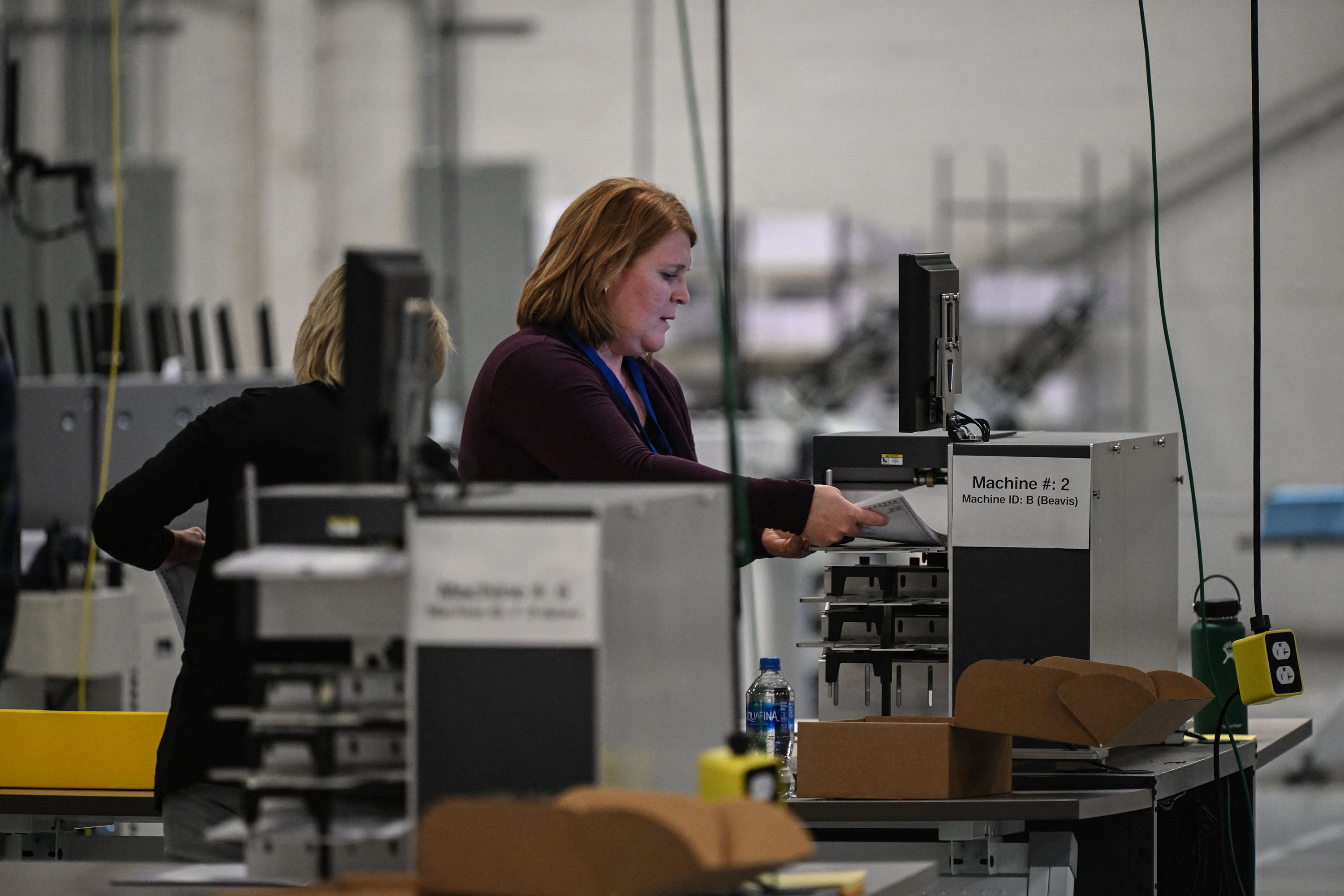 A person with short red hair stands at a ballot machine with other machines in the foreground and background.