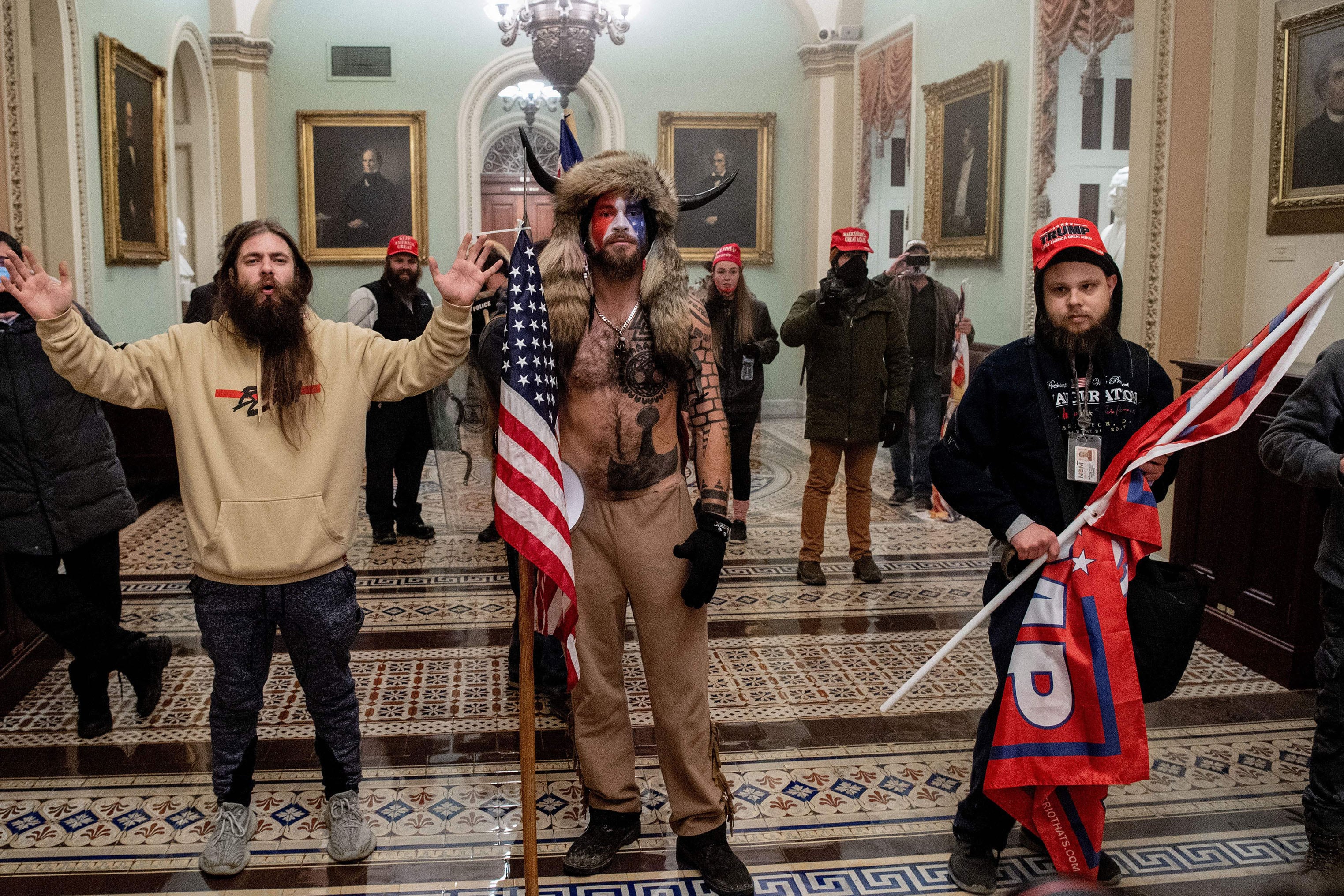 Three people stand in a row inside of teh US Capitol. Two of the three men are holding a flag, the two men on each side are wearing sweatshirts and the man in the middle is wearing a fur hat with horns coming out of the side and no shirt.