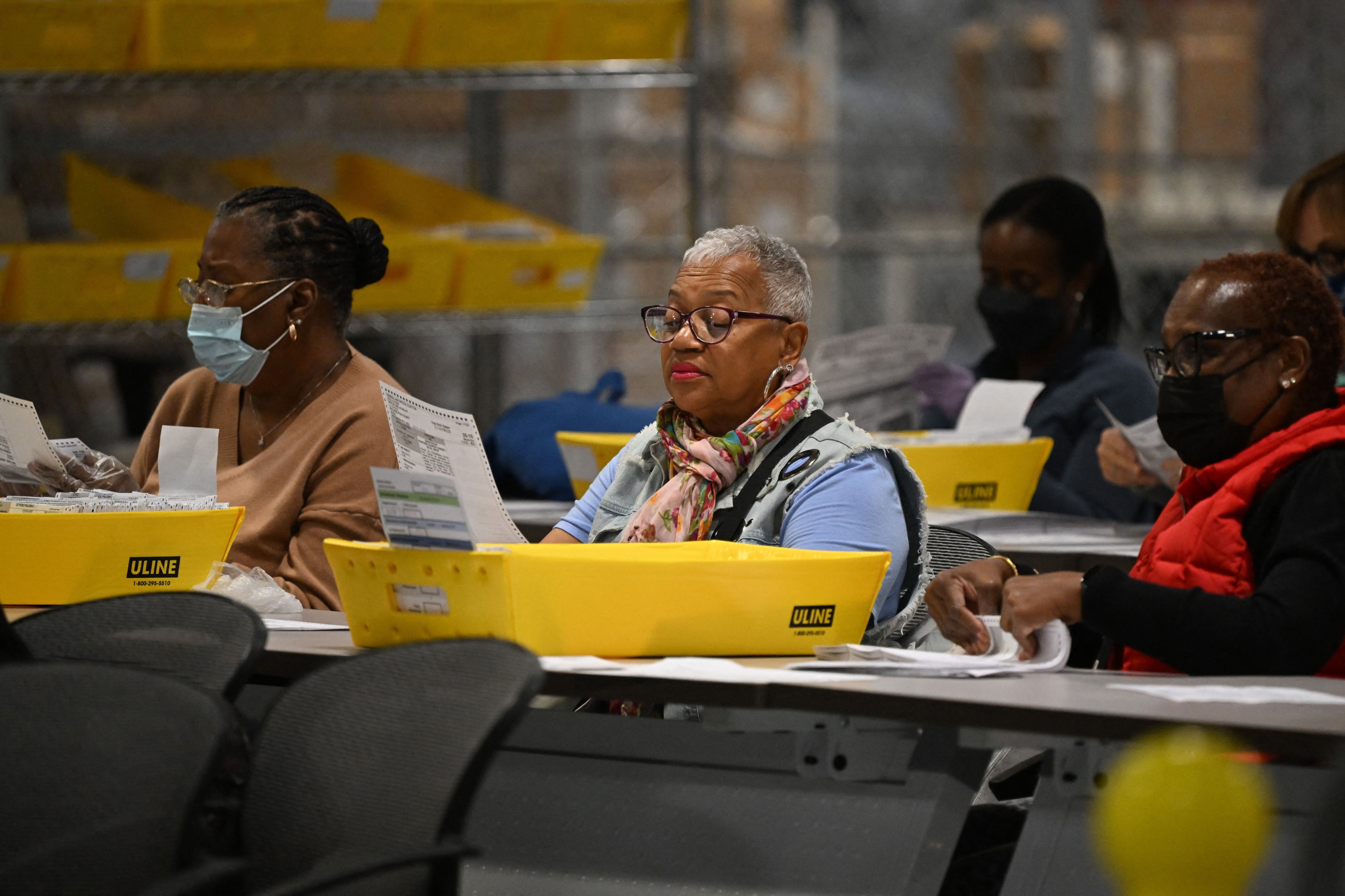 A woman with a scarf and glasses sits with other women processing ballots in a warehouse.