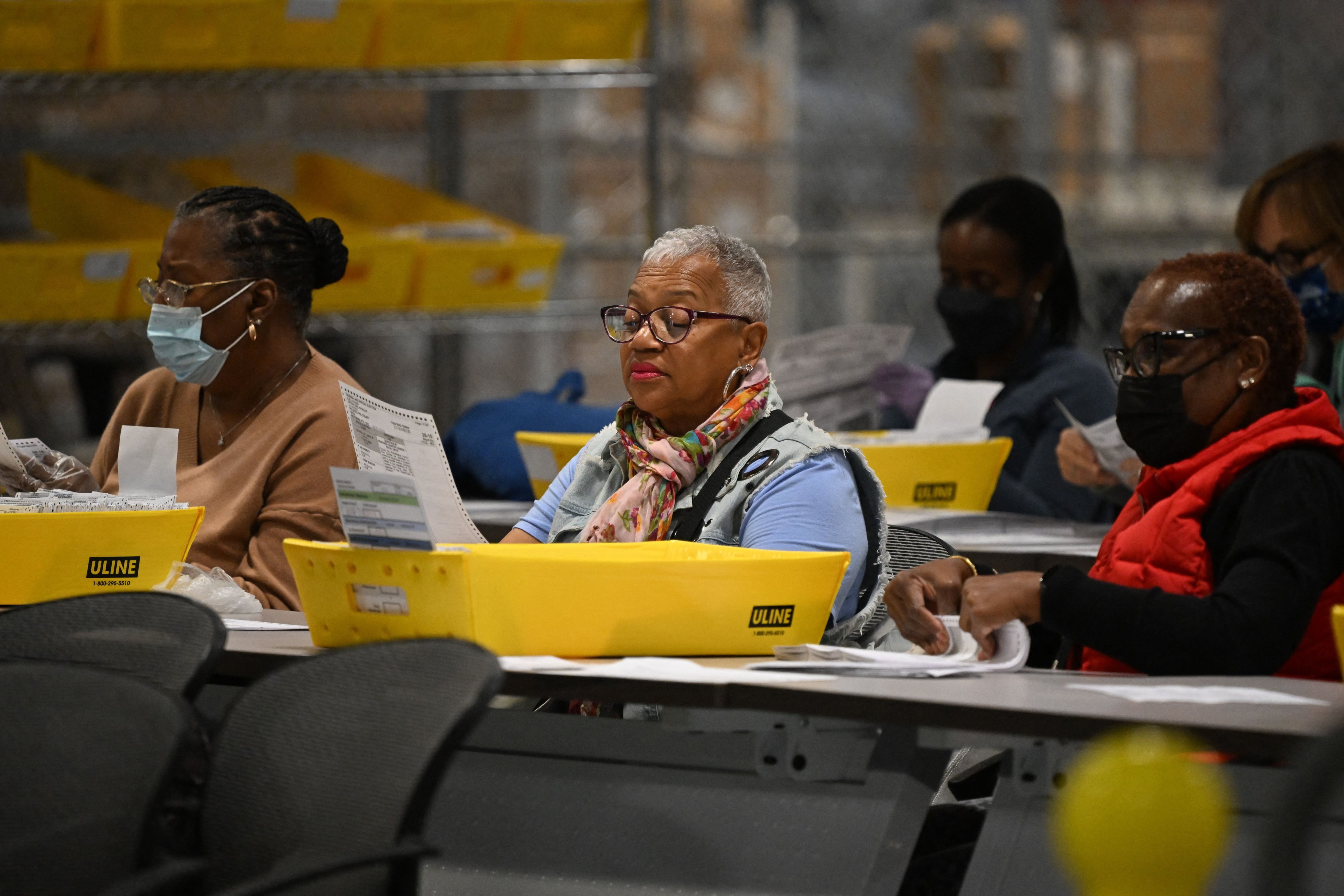 A woman with a scarf and glasses sits with other women processing ballots in a warehouse.