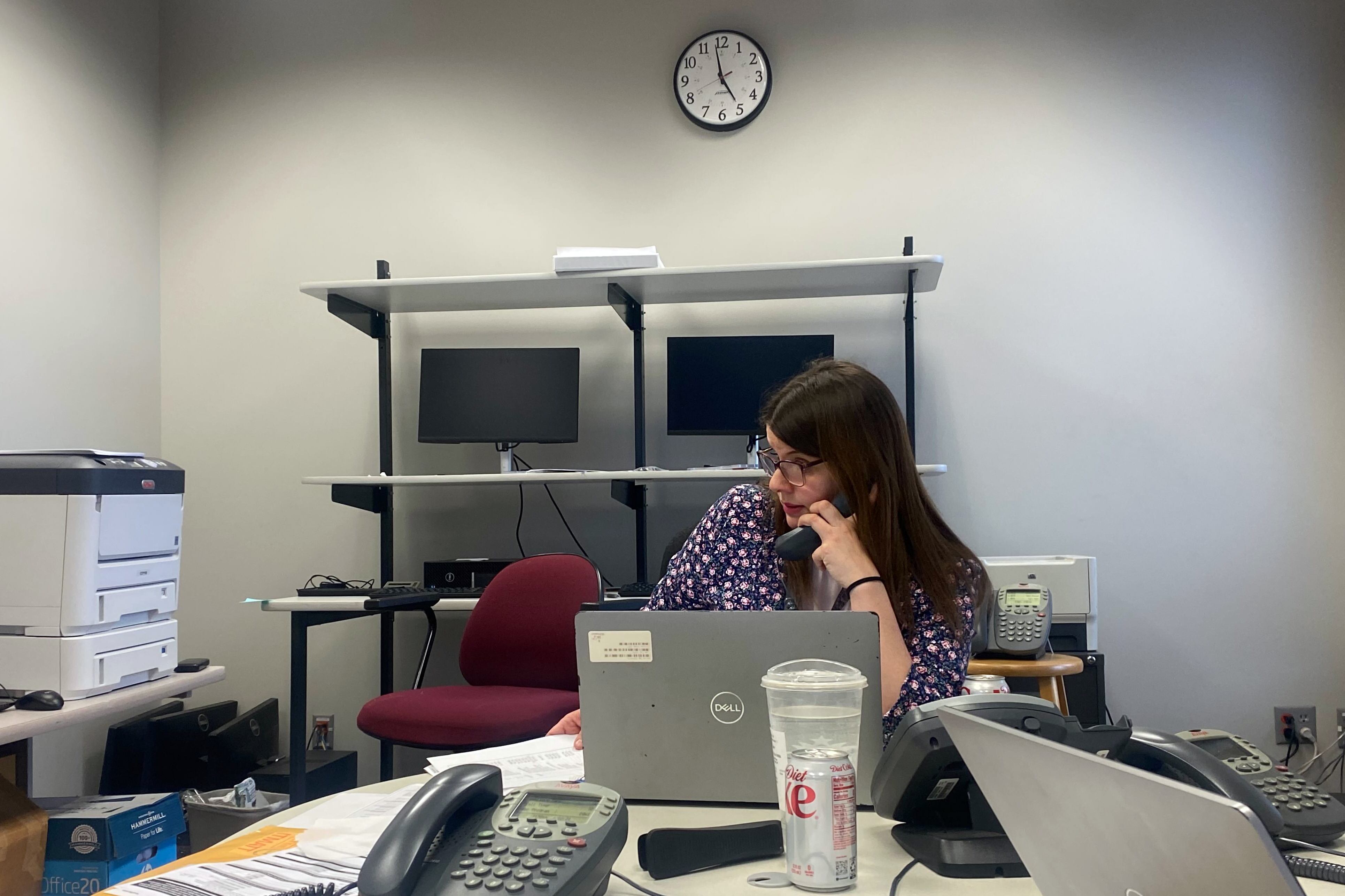 A woman with long brown hair and wearing a purple and pink blouse works at a desk typing on a laptop in an office.