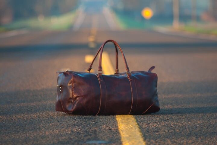A brown leather suitcase sitting on the yellow hashed line of a sunny countryside highway