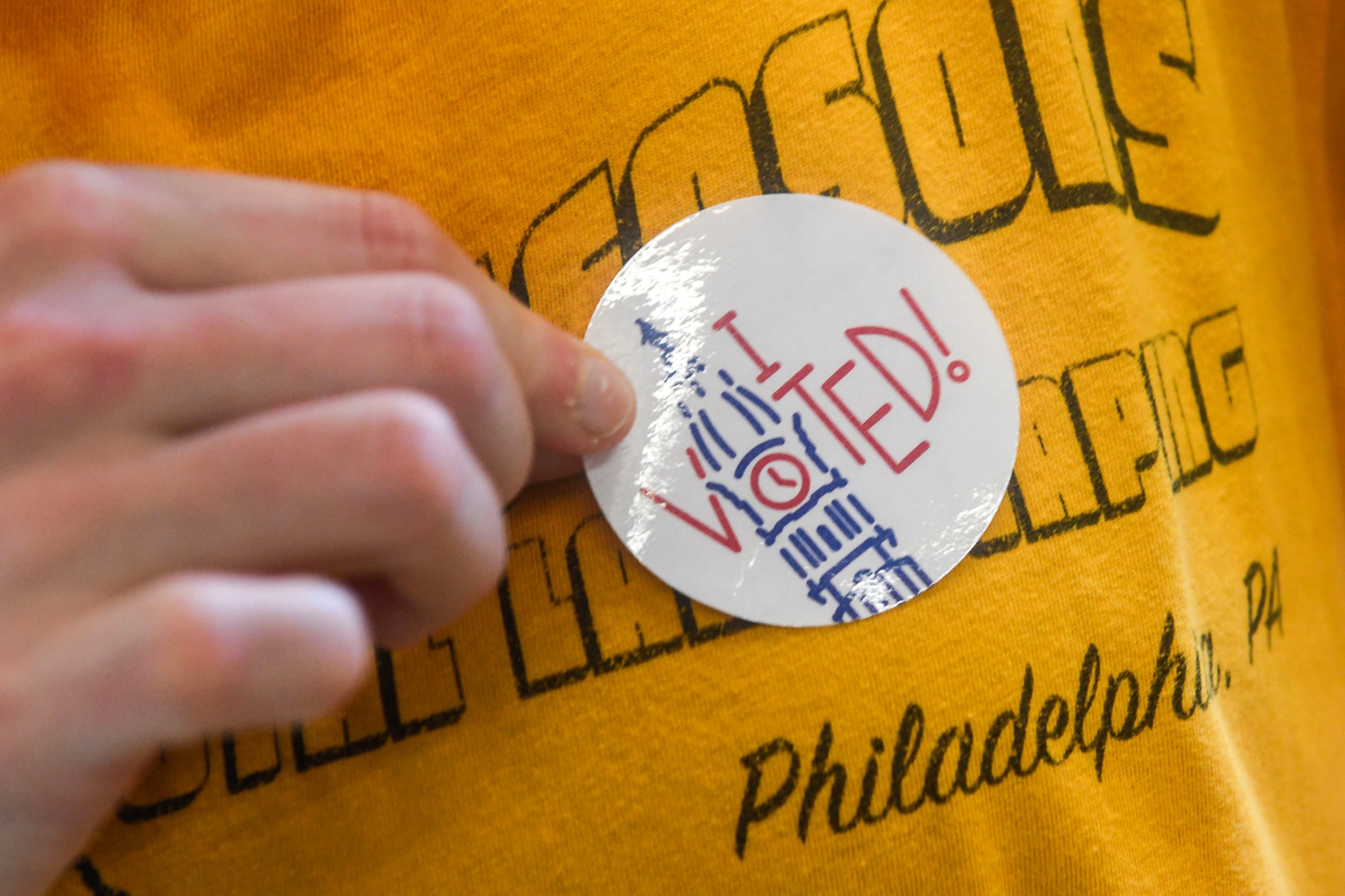 A close up photograph of a person placing an "I voted" sticker on their bright yellow t-shirt.