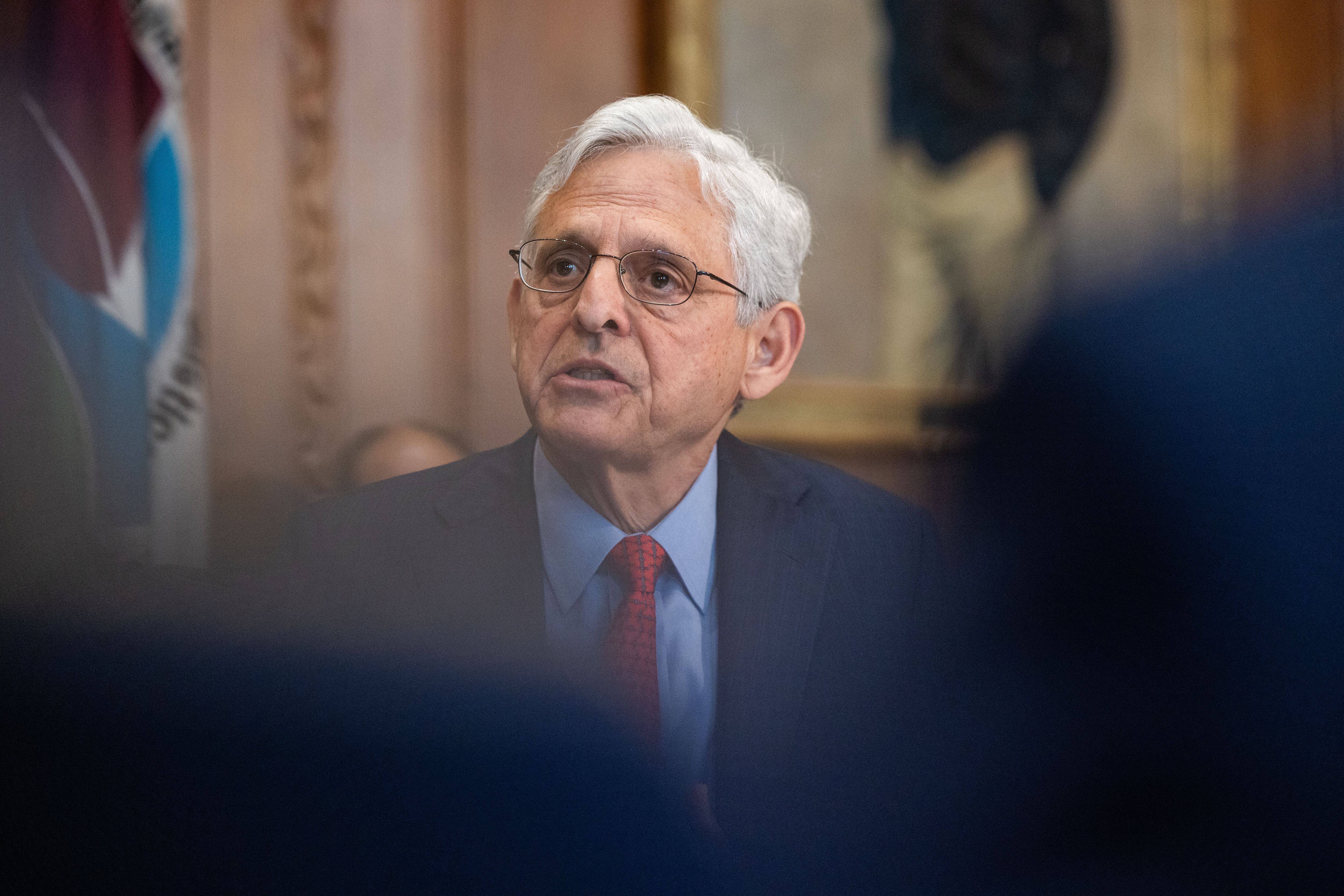 A man with white hair and wearing glasses and a dark suit speaks in a room.