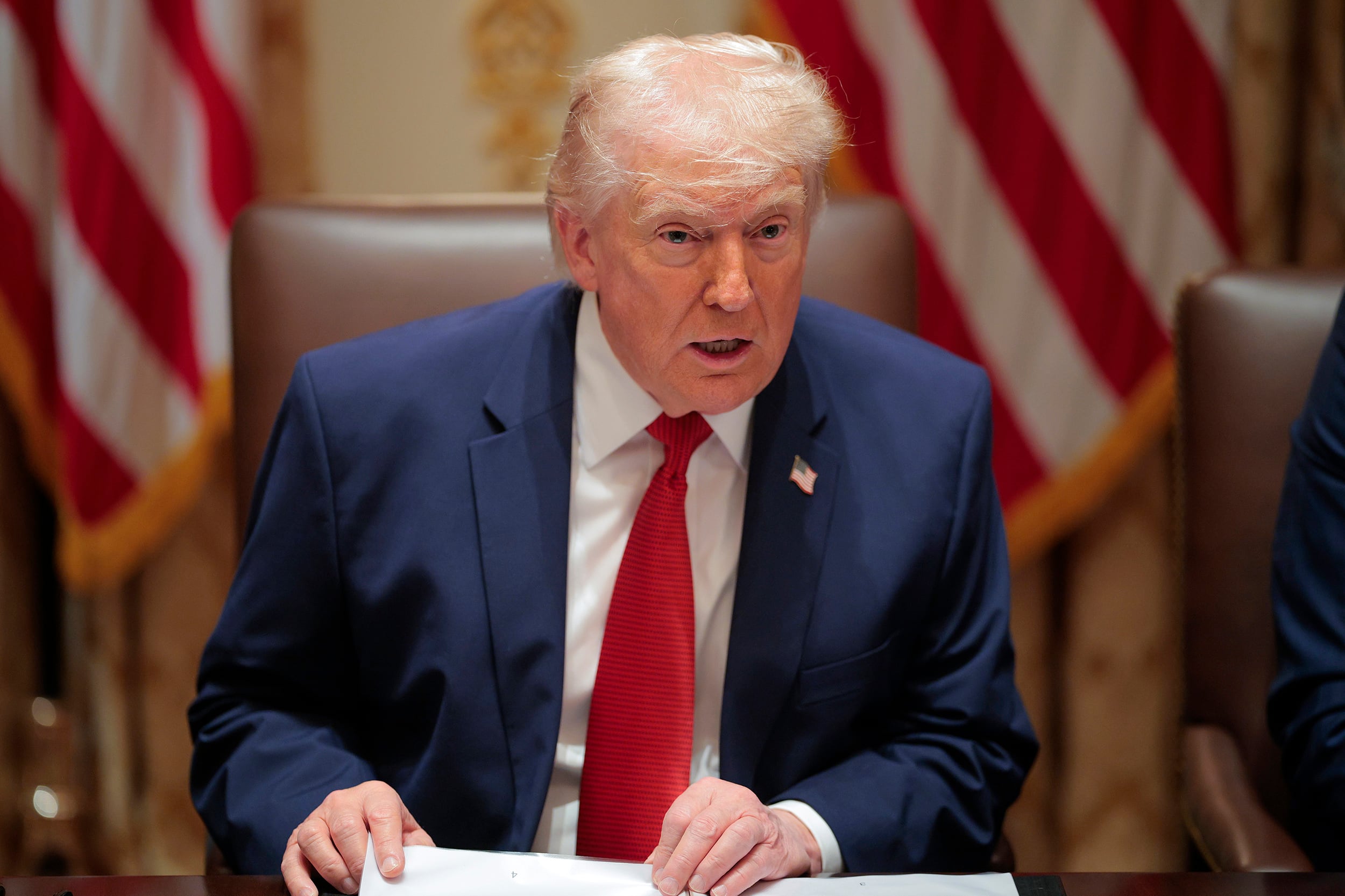 A photograph of President Donald Trump, a white man with white hair wearing a blue suit jacket and a red tie sits at a table speaking with two American flags in the background.