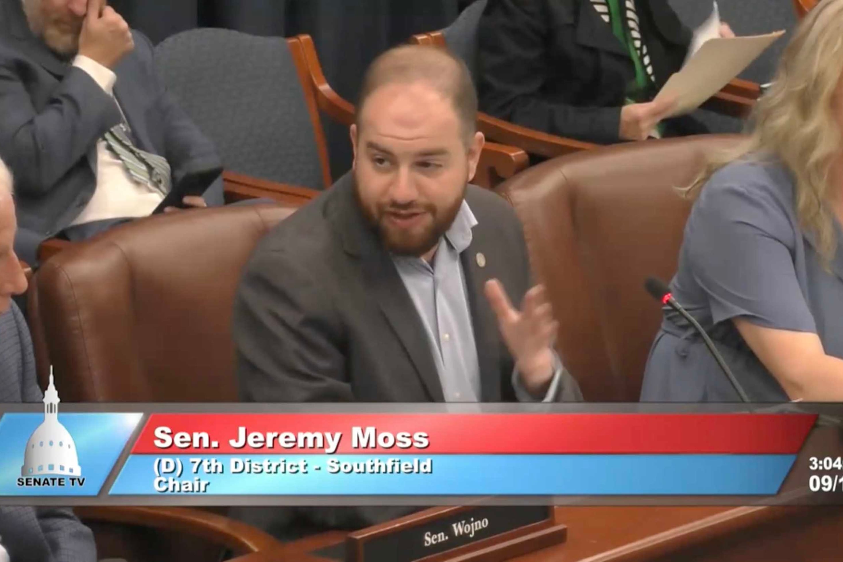 A screen grab of a livestream feed of three adults in suits sitting at a table.