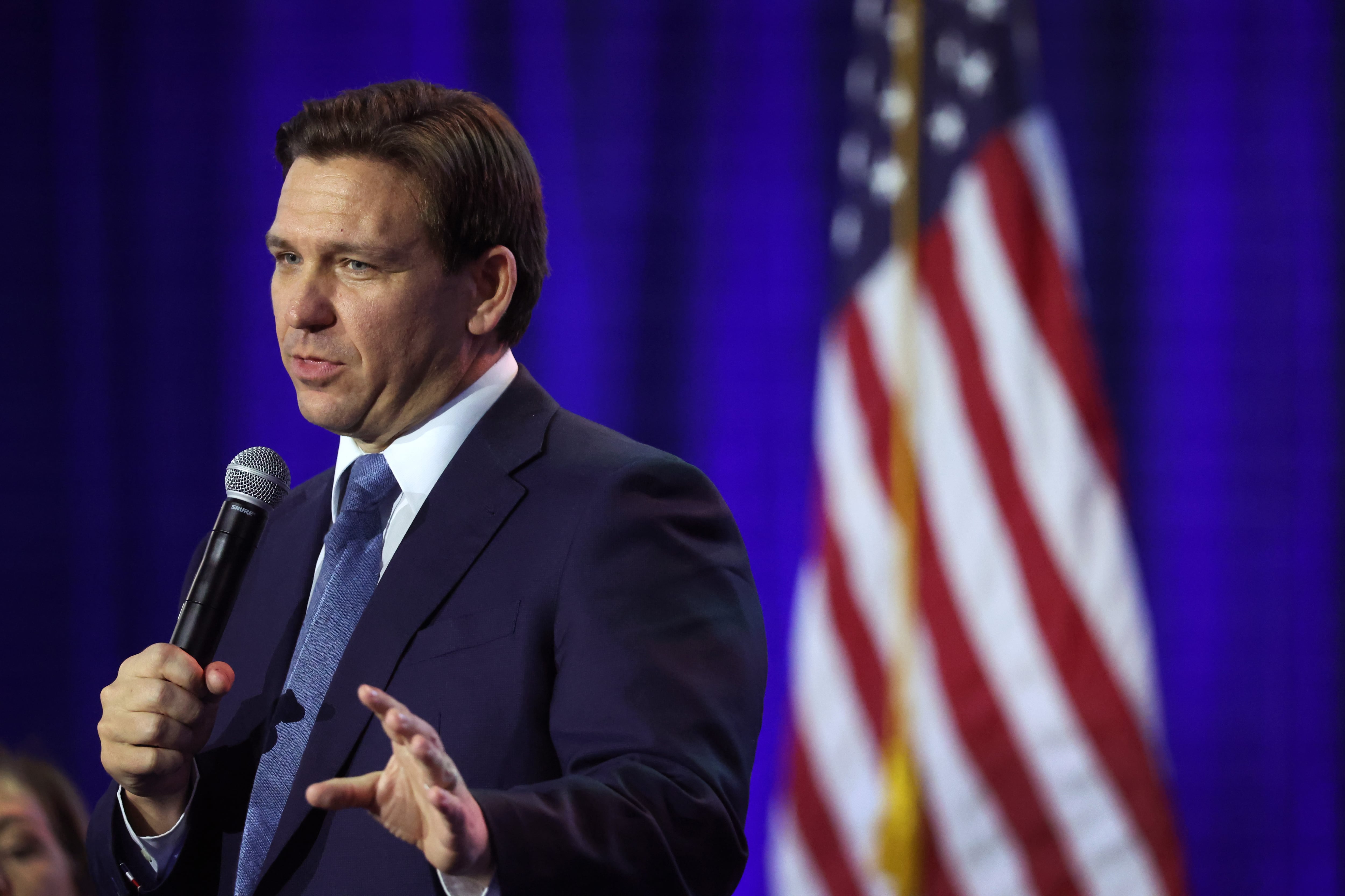 A man gestures while speaking into a microphone, with an American flag behind him.