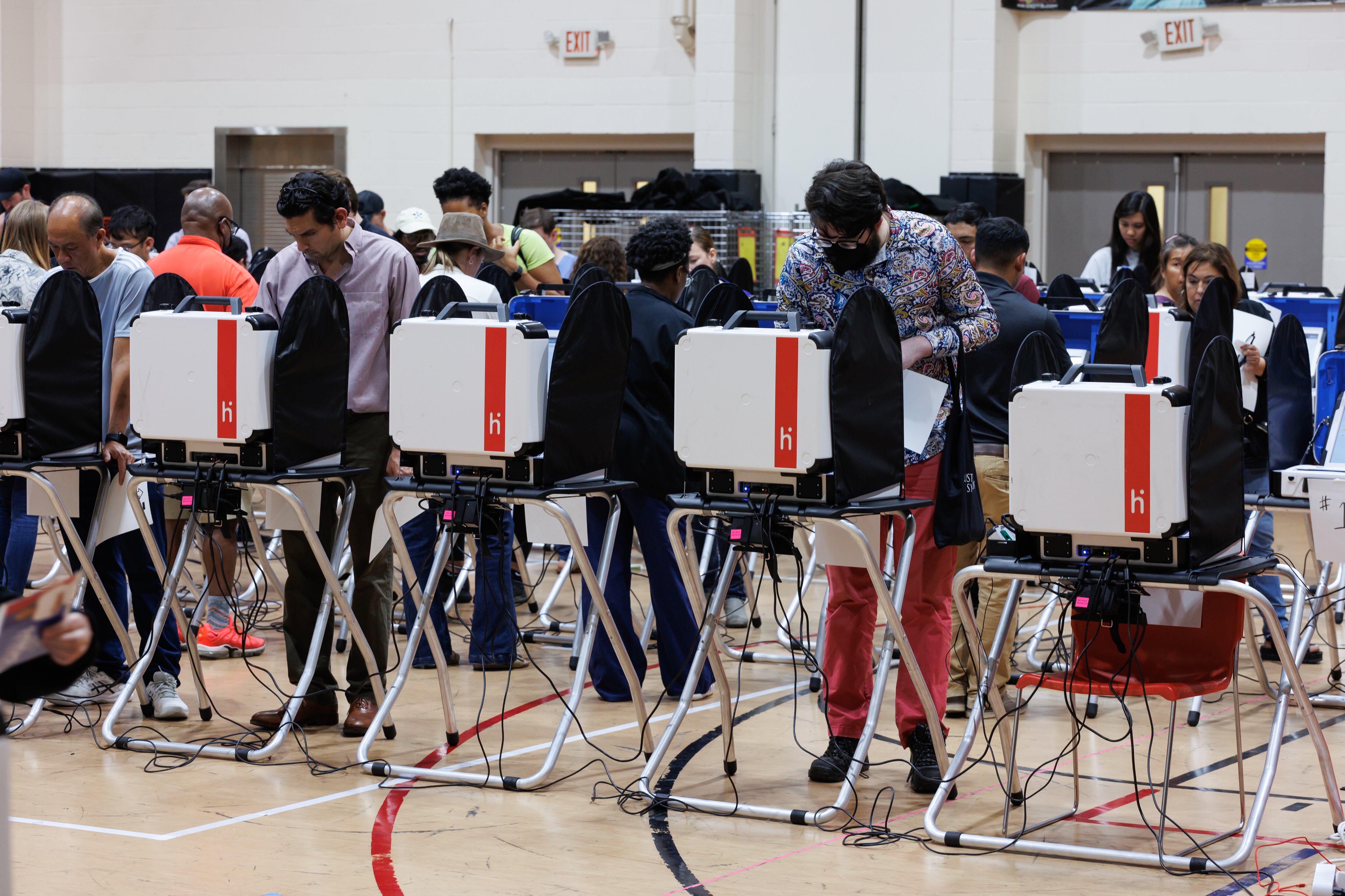 A line of voting machines in a gymnasium, with people standing at each