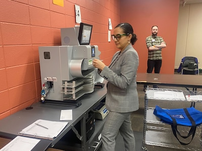 A woman wearing a gray suit works at a machine in a room with a man standing near an orange brick wall in the background.