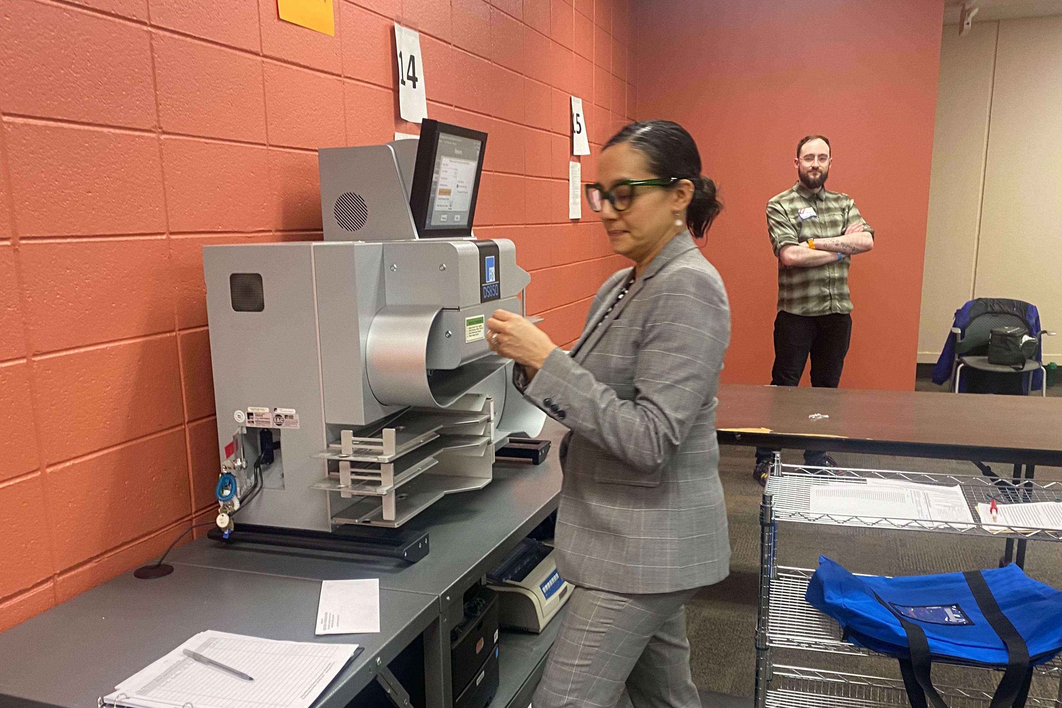 A woman wearing a gray suit works at a machine in a room with a man standing near an orange brick wall in the background.