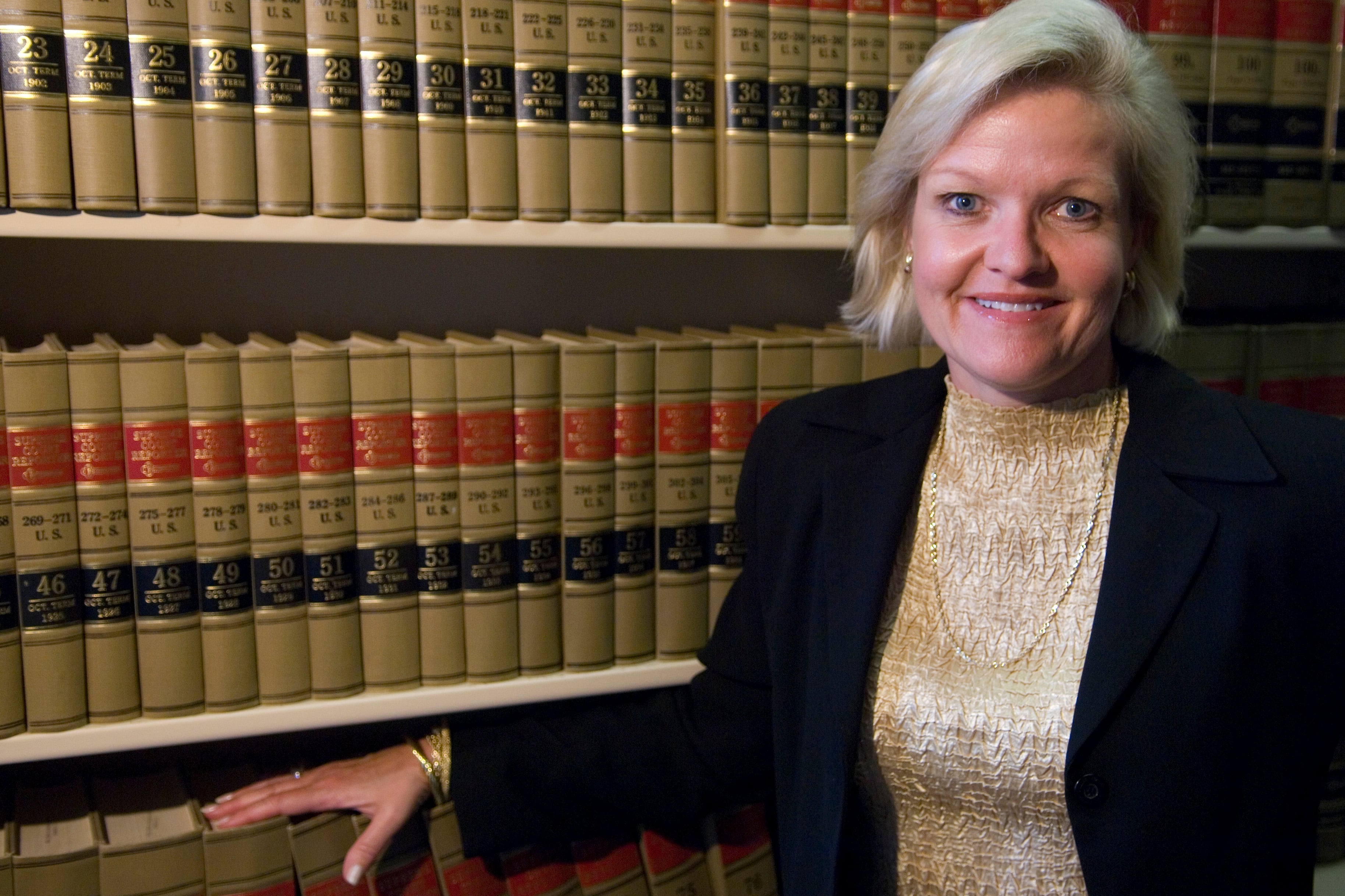 Cleta Mitchell wearing a suit jacket and gold shirt standing before a tall shelf of law books.