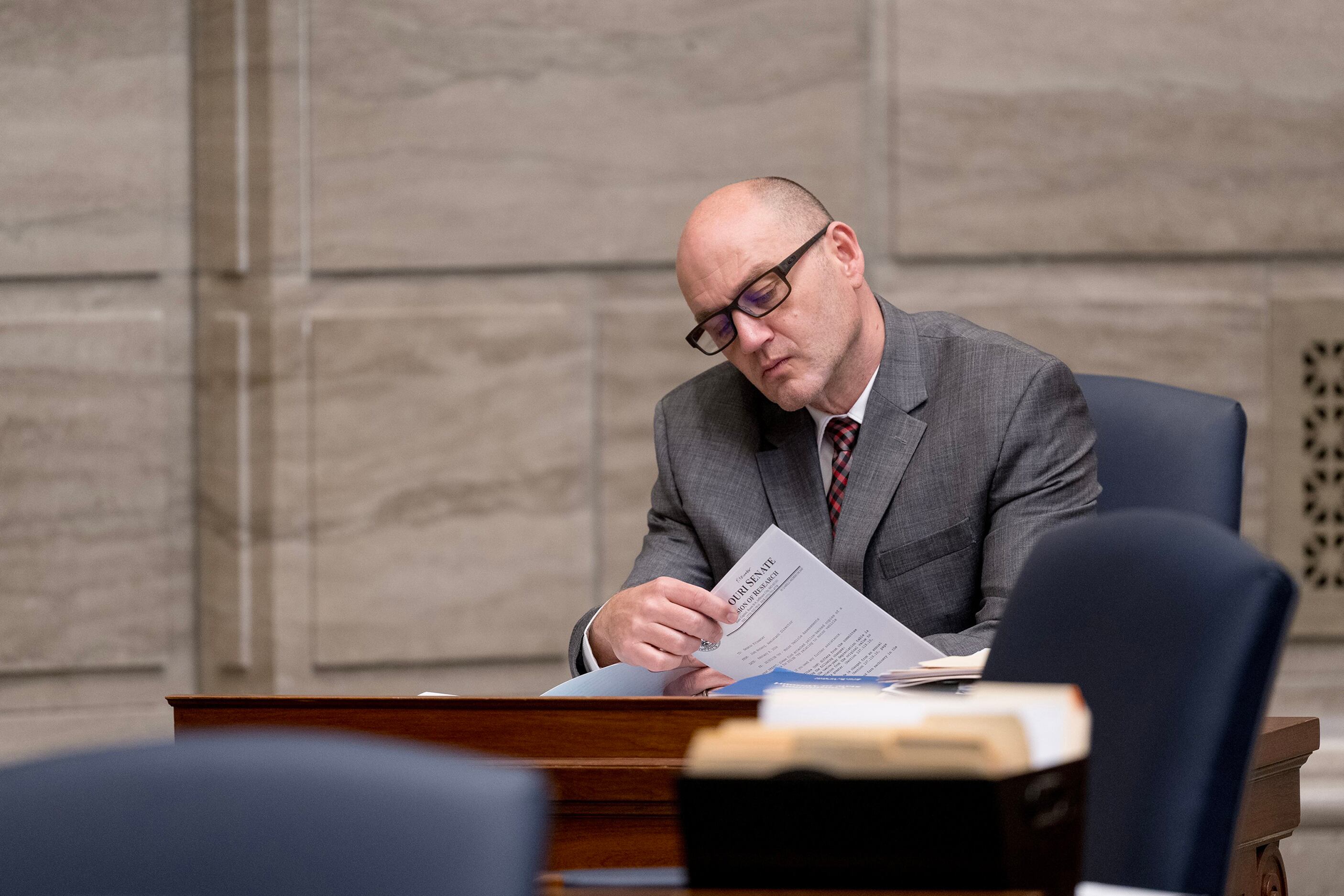 A man with glasses and wearing a grey suit sits at a wooden desk looking through papers with a stone wall in the background.