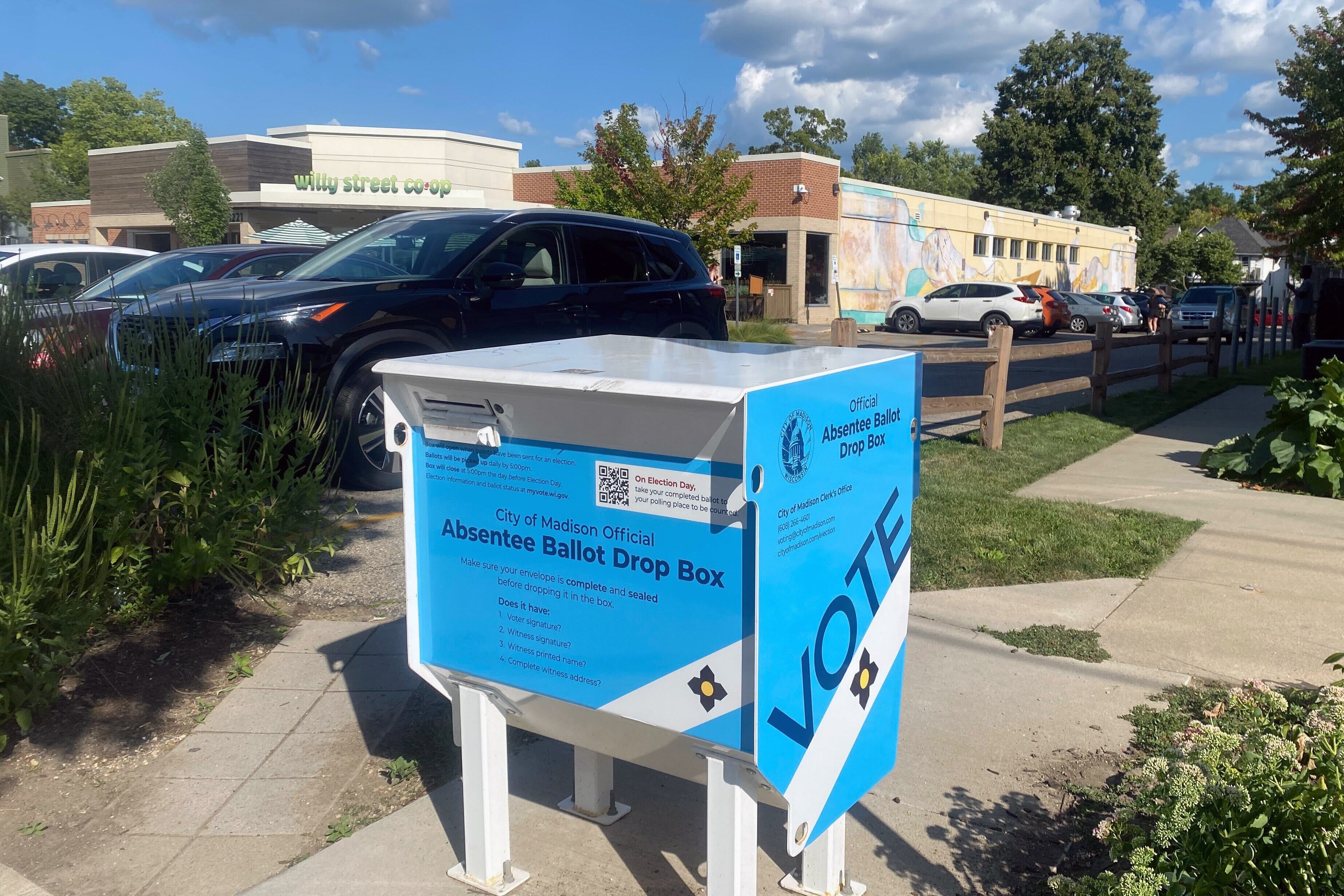A blue and white large box stands on concrete outside with cars and a building in the background.