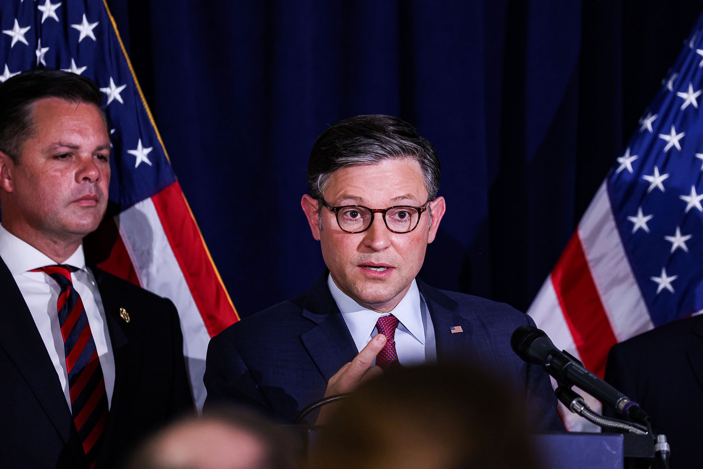 Two men in suits stand next to each other behind a microphone and in front of a dark background with two American flags.