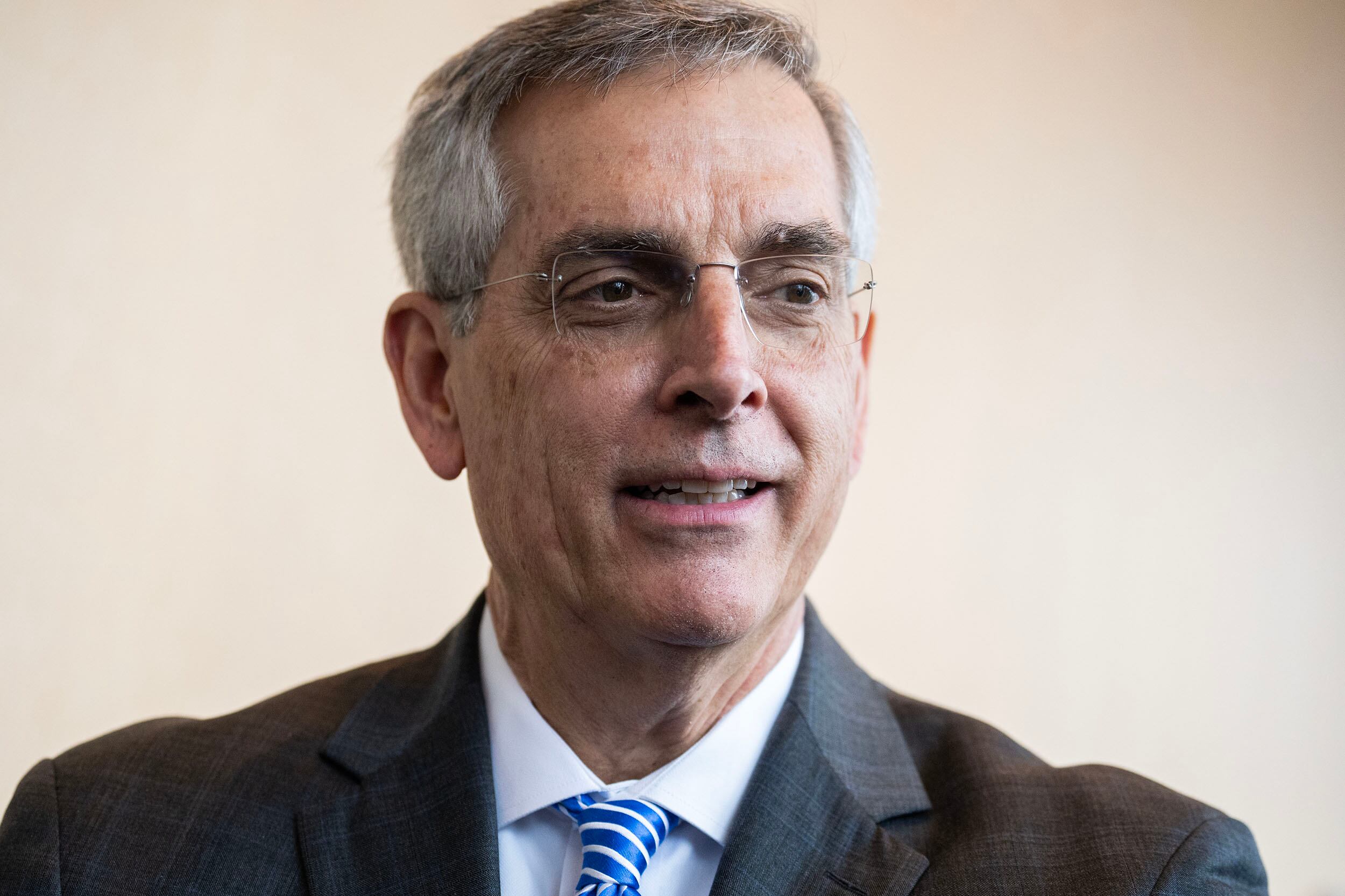 A man with glasses and wearing a dark suit looks away from the camera while standing in front of a tan wall.