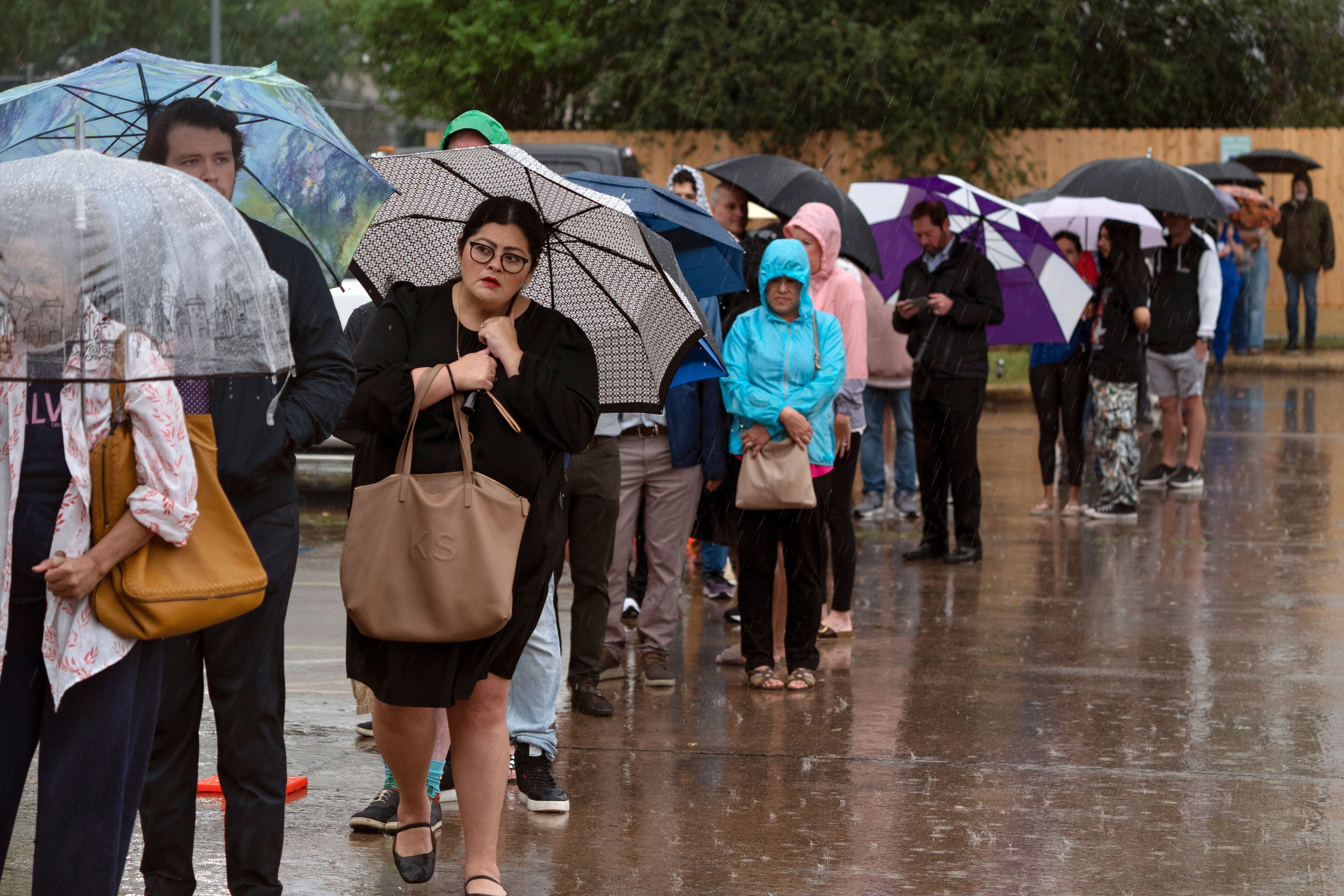 A long line of people stand in the rain, some with umbrellas.