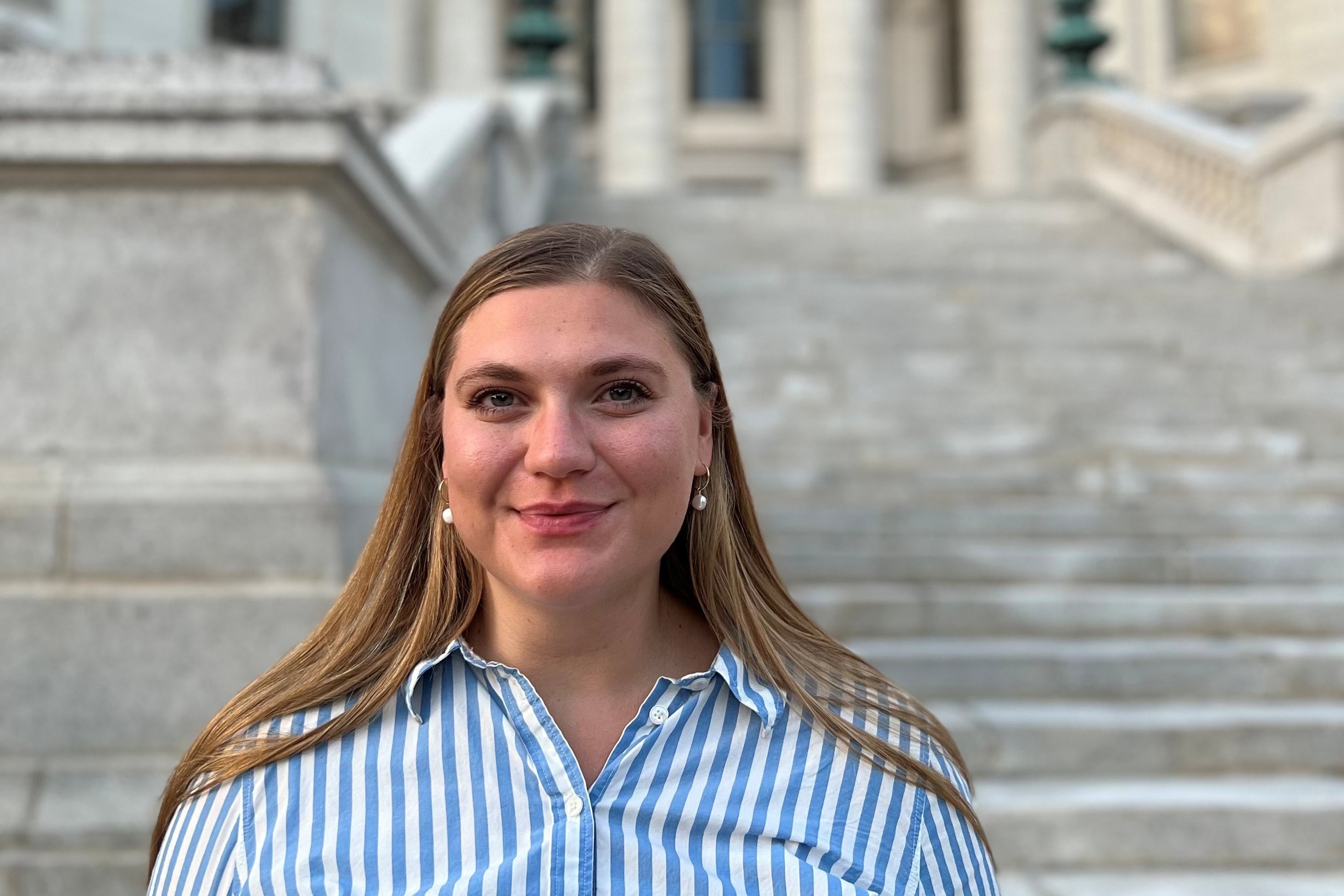 A photograph of a white woman with long blond hair and wearing a blue and white striped blouse smiling while standing in front of a large stone building.