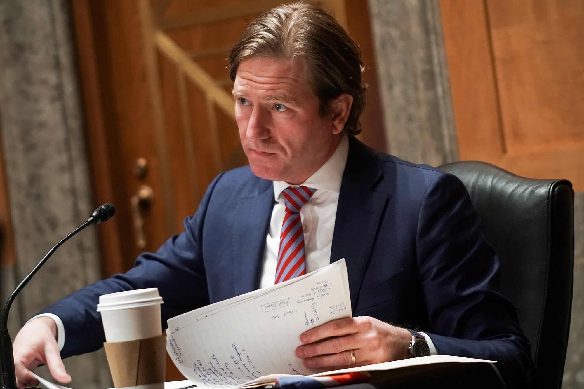 A white man in a dark suit and wearing a red tie looks up from his papers in a large room.