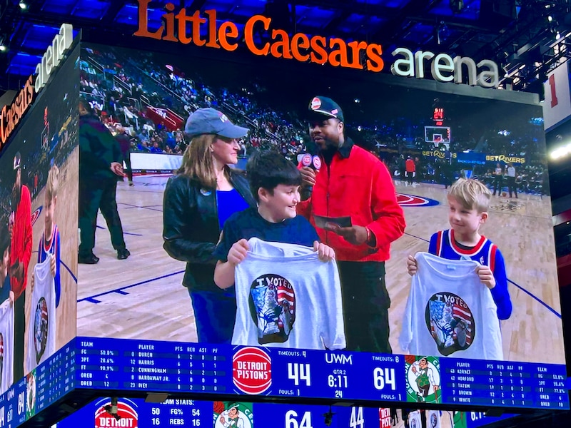 A photograph of a large TV screen in a basketball arena that shows a white woman standing next to two kids who are holding up t-shirts and a Black man who is interviewing her. The words "Little Caesars Arena" are on the top of the screen.