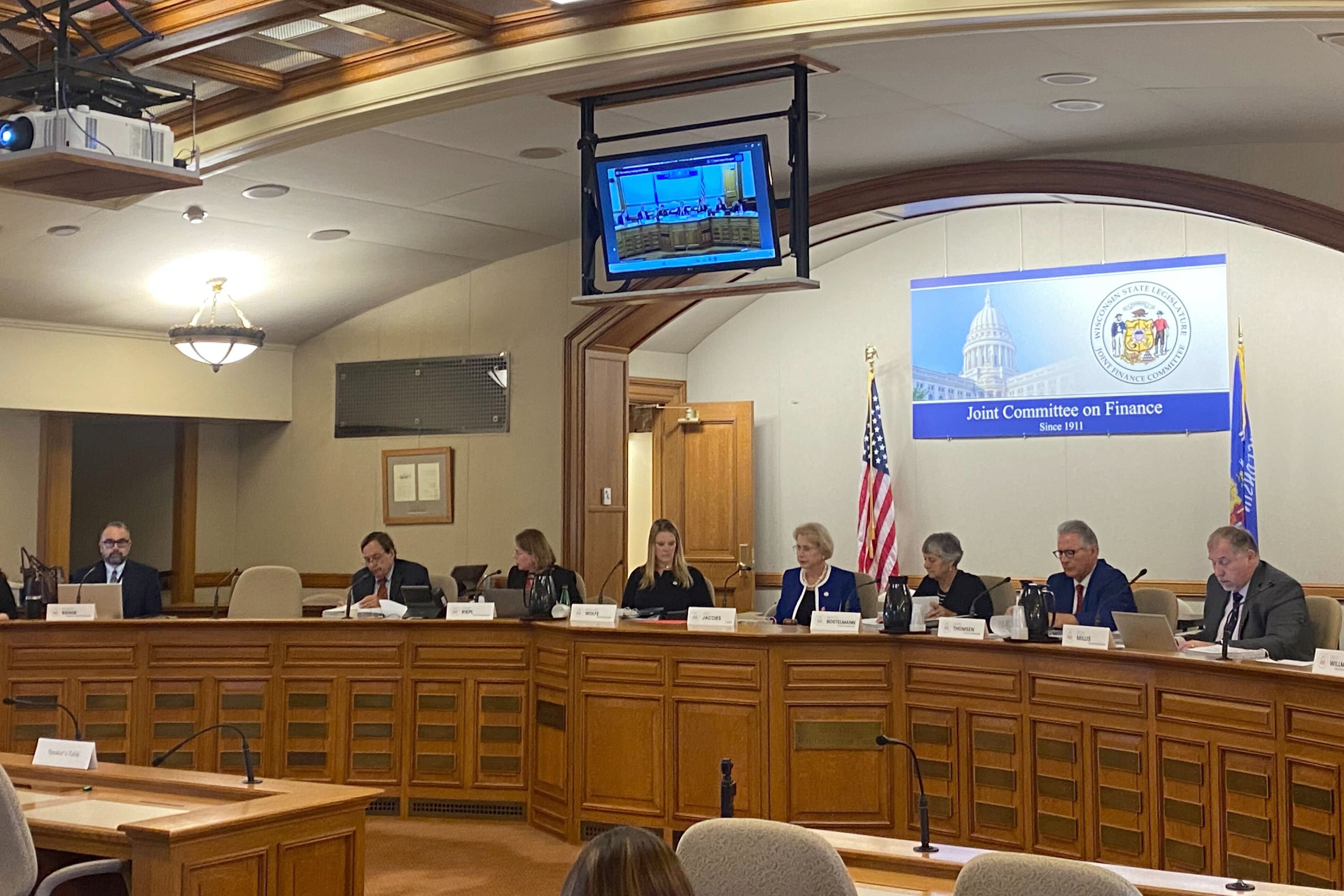 A photograph of a group of people sitting at a long wooden table with a TV hanging from the ceiling.