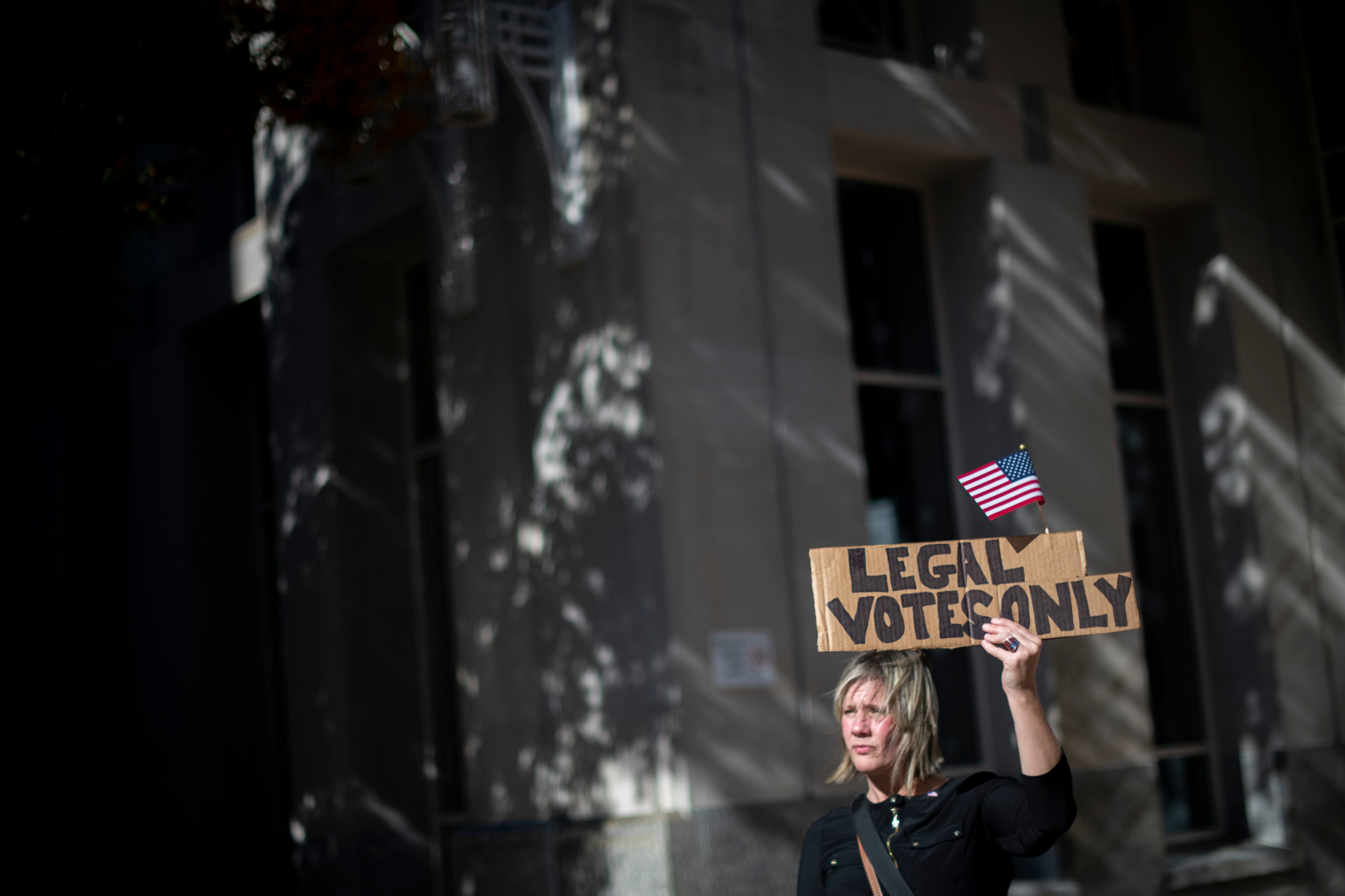 A woman holds a sign that reads “Legal Votes Only” outside of a building. Windows reflect the columns of the building’s facade across the frame.