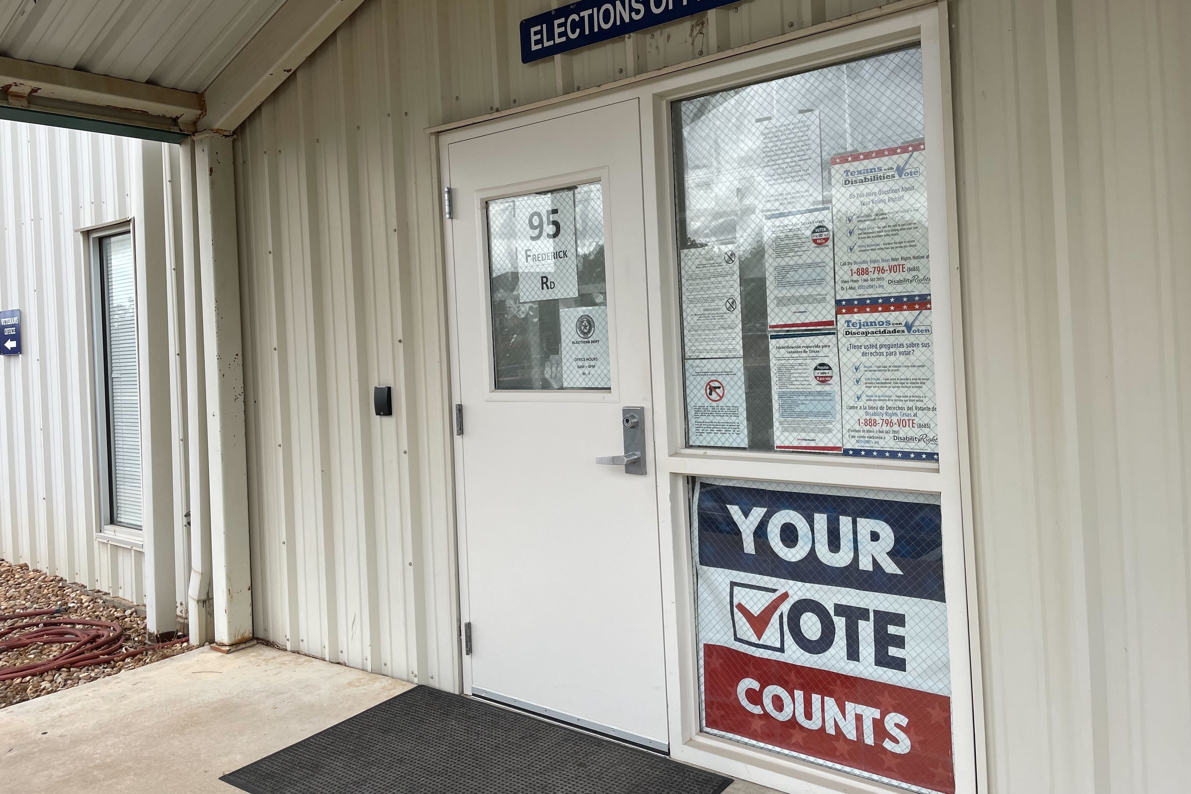 A door and window of a white building with signs reading “Your Vote Counts” and “Elections Office”
