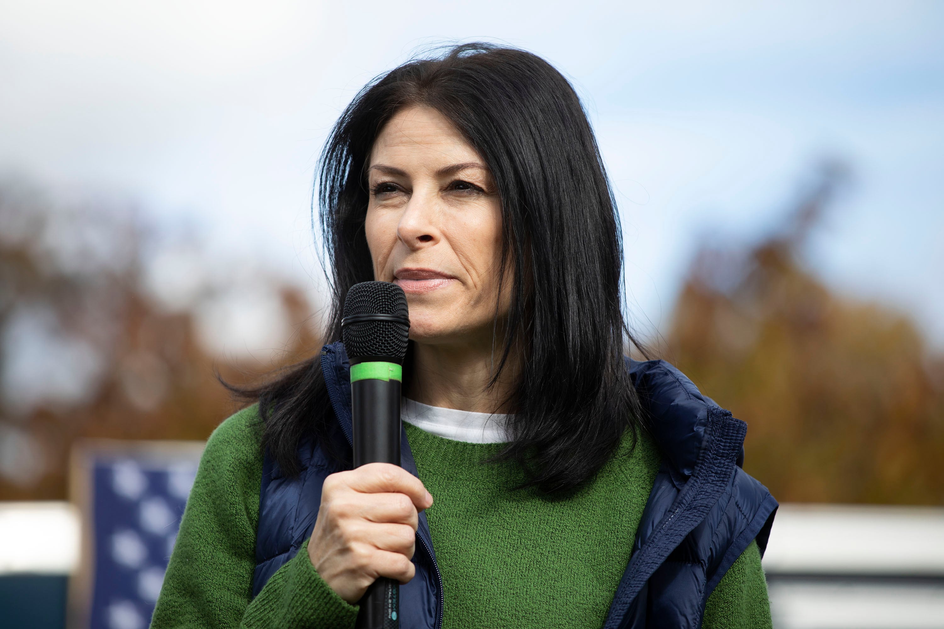 A photograph of a white woman with long dark hair holding a microphone while standing outside.