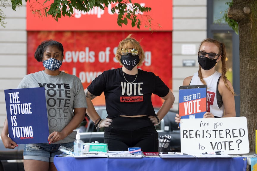 Three young women standing at an outdoor table wearing T-shirts reading “Vote,” behind a sign reading “Are you registered to vote?” 
