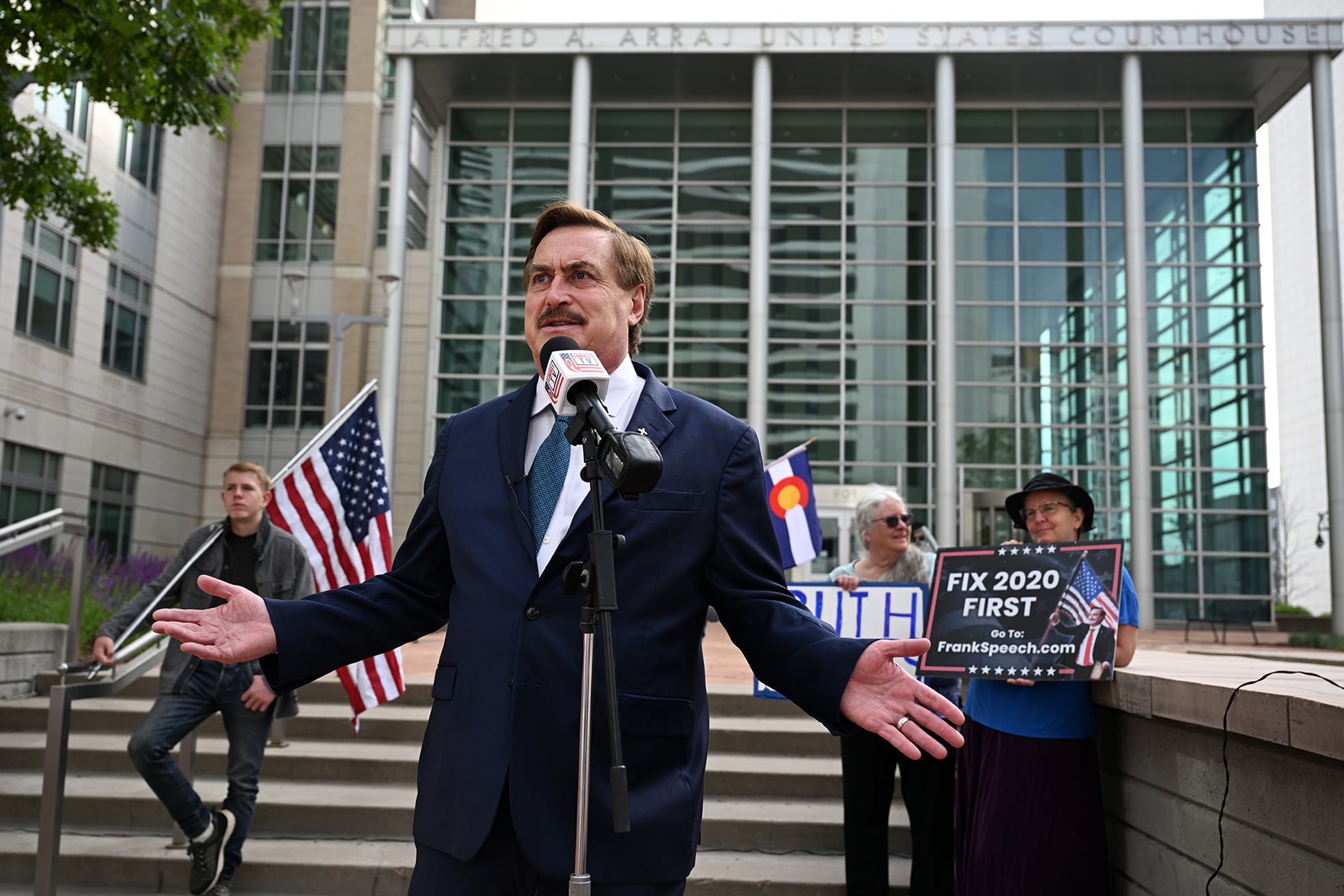 A photo of a white man in a dark suit with his arms out, speaks from a microphone outside of a large glass building with a small group of people in the background holding up signs and an American flag.