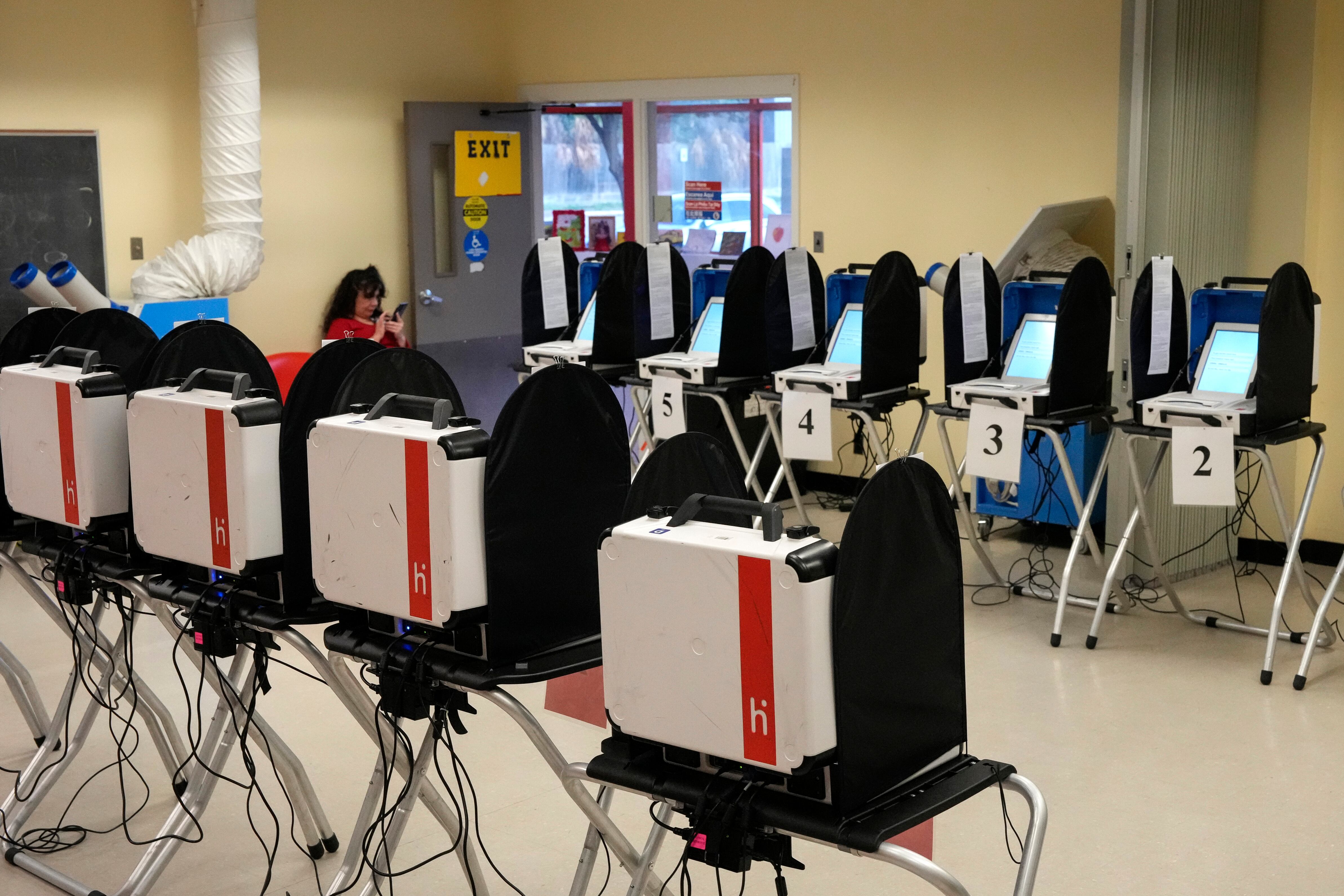 Two rows of voting machines are in two lines in a room with one person sitting on a chair on the far left.
