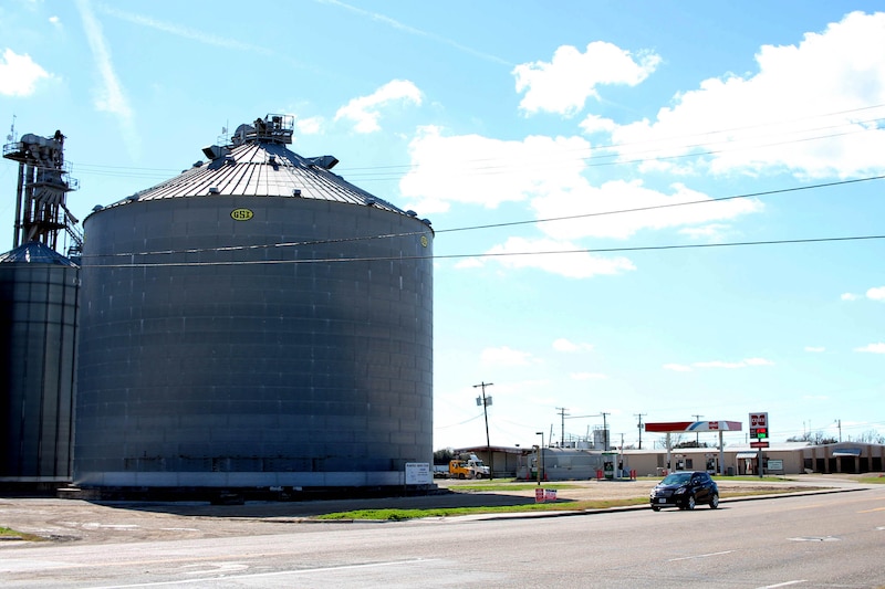 A large grey grain silo sits next a a road with a blue car and a gas station in the background.