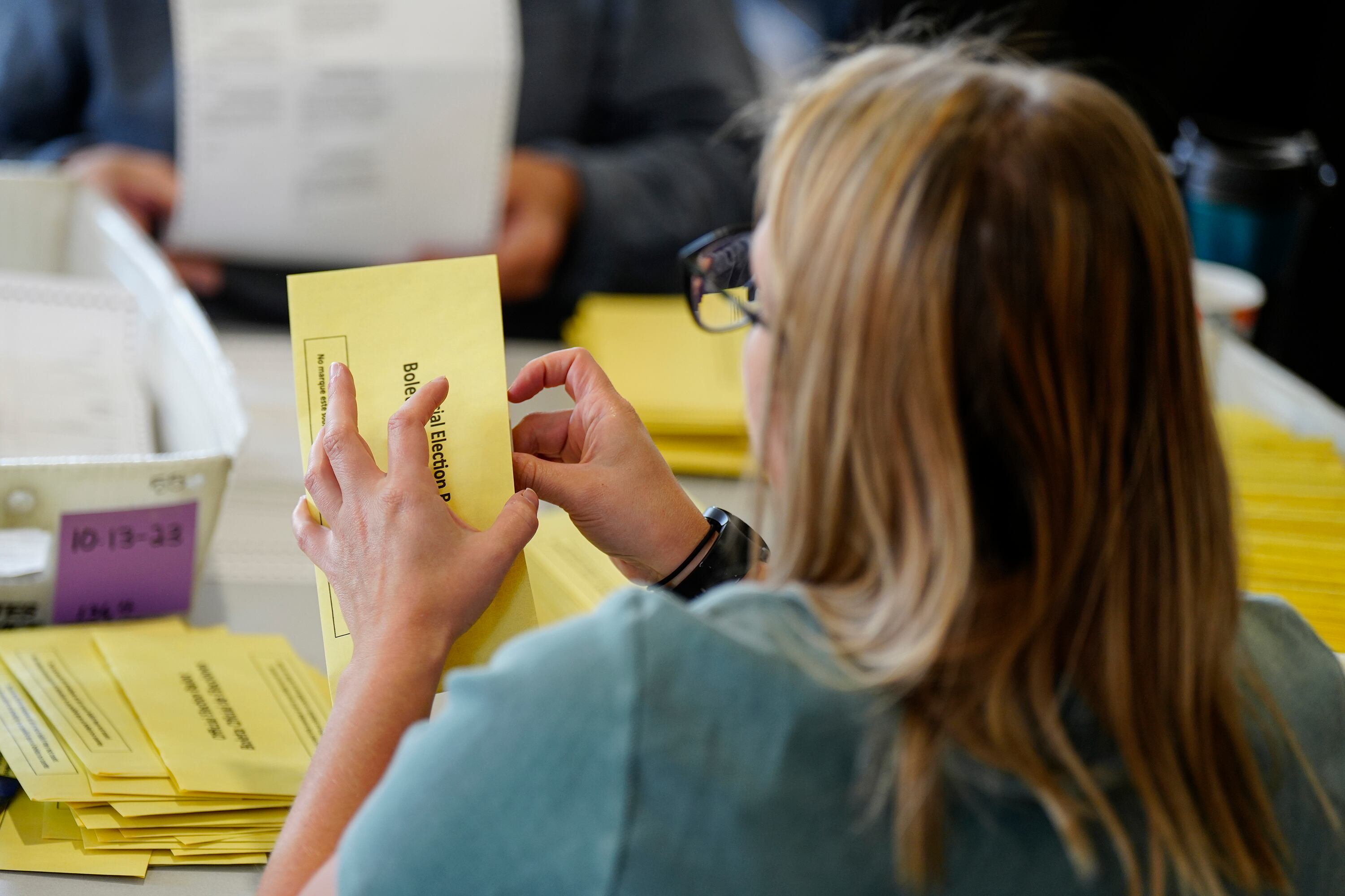 A person with long blonde hair and wearing a blue shirt holds a yellow ballot envelope with stacks of yellow envelopes on the table in the background.