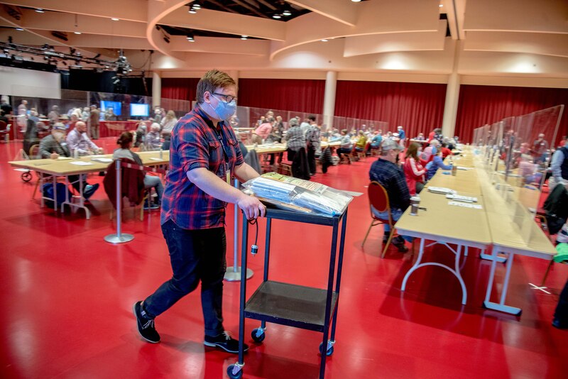 A person wearing a mask and a blue and red striped shirt pushing a cart next to rows of long tables with people sitting.