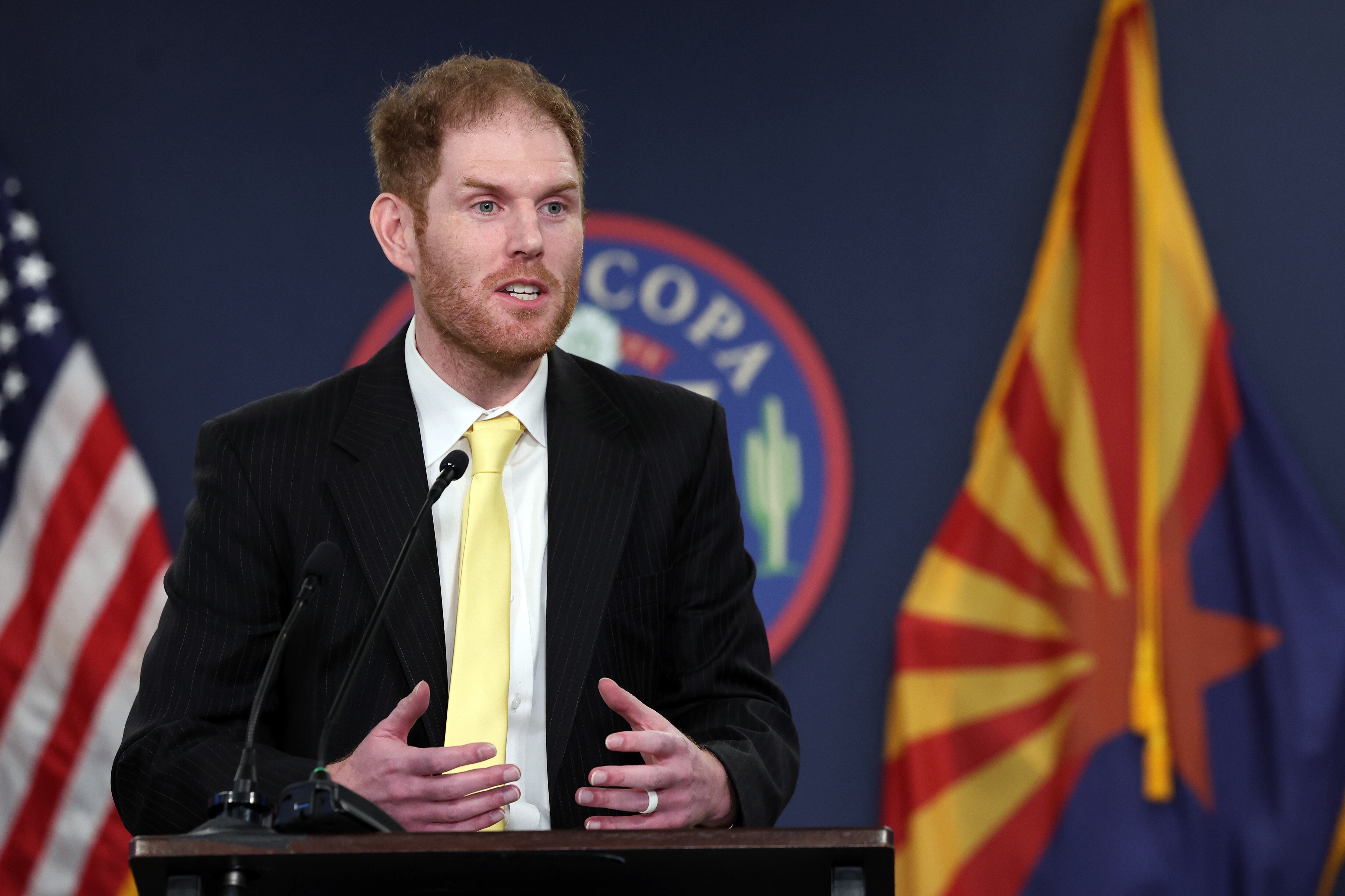 A man wearing a dark suit and a yellow tie speaks from behind a podium with a microphone and in front of a dark background with an American and Arizona flag.