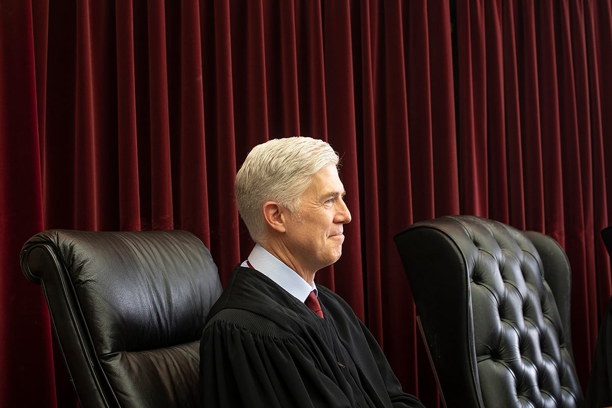 A man with white hair and wearing a black robe sits on a leather chair with a red velvet curtain in the background.