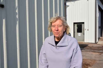 A photograph of a white woman in a sweater posing for a portrait outside of a building on a sunny day.