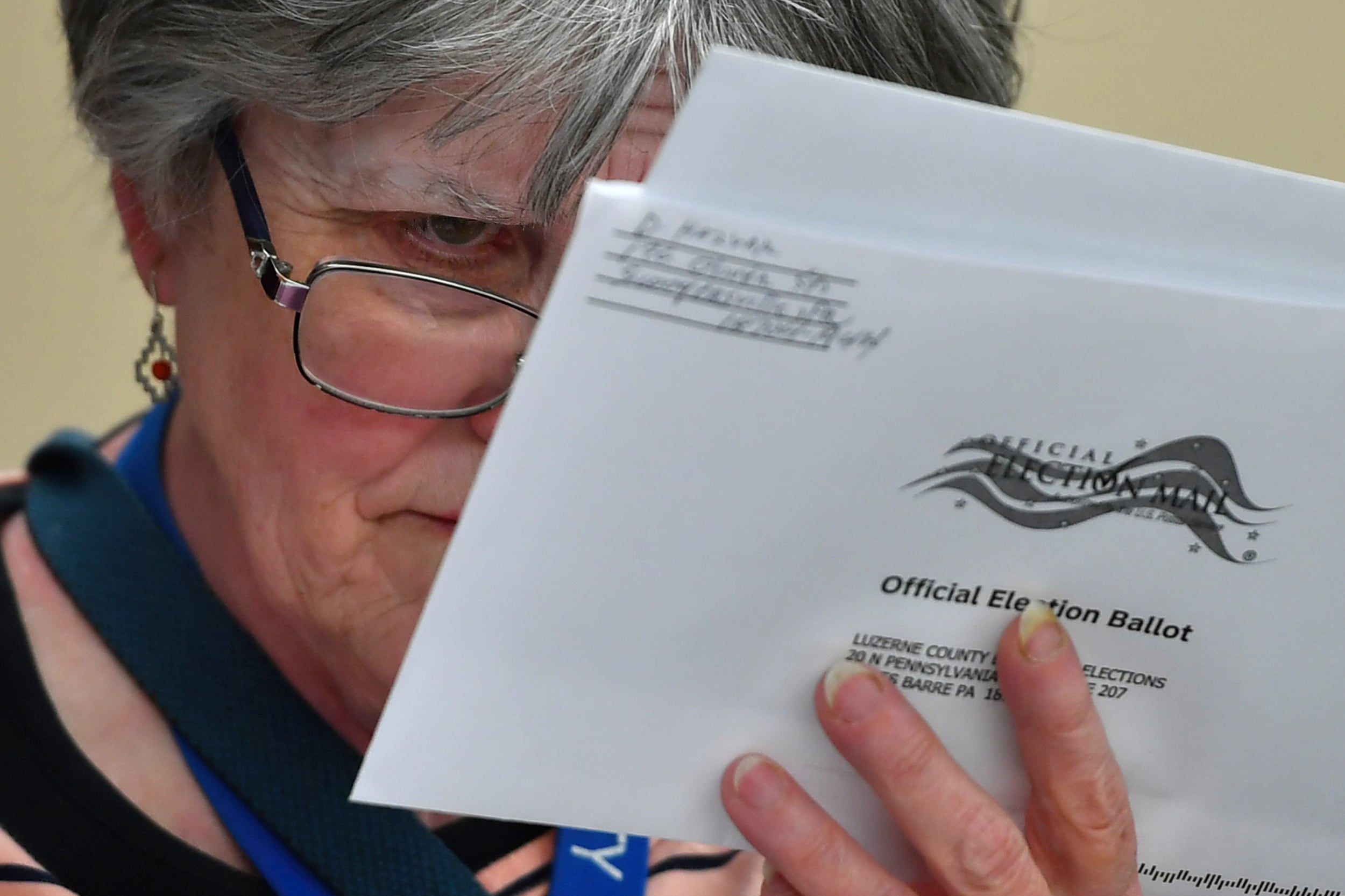A woman with glasses and grey hair holds a white mail-in paper ballot close to her face to inspect the handwriting.