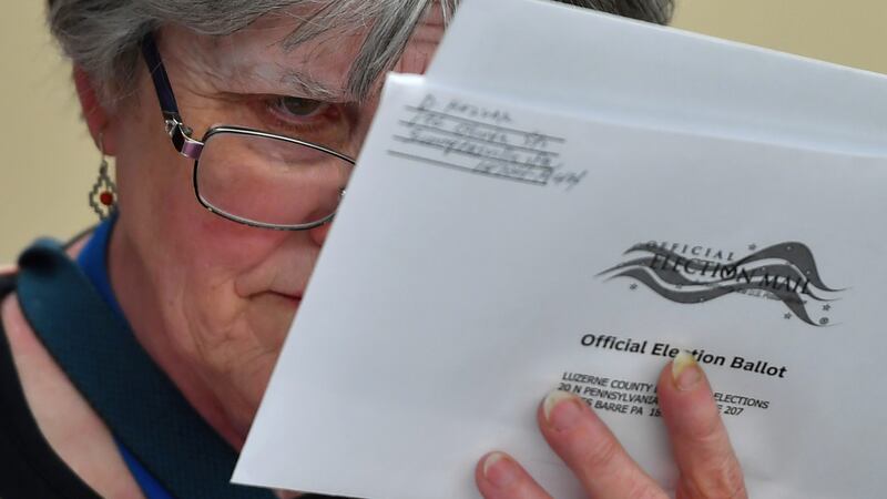 A woman with glasses and grey hair holds a white mail-in paper ballot close to her face to inspect the handwriting.