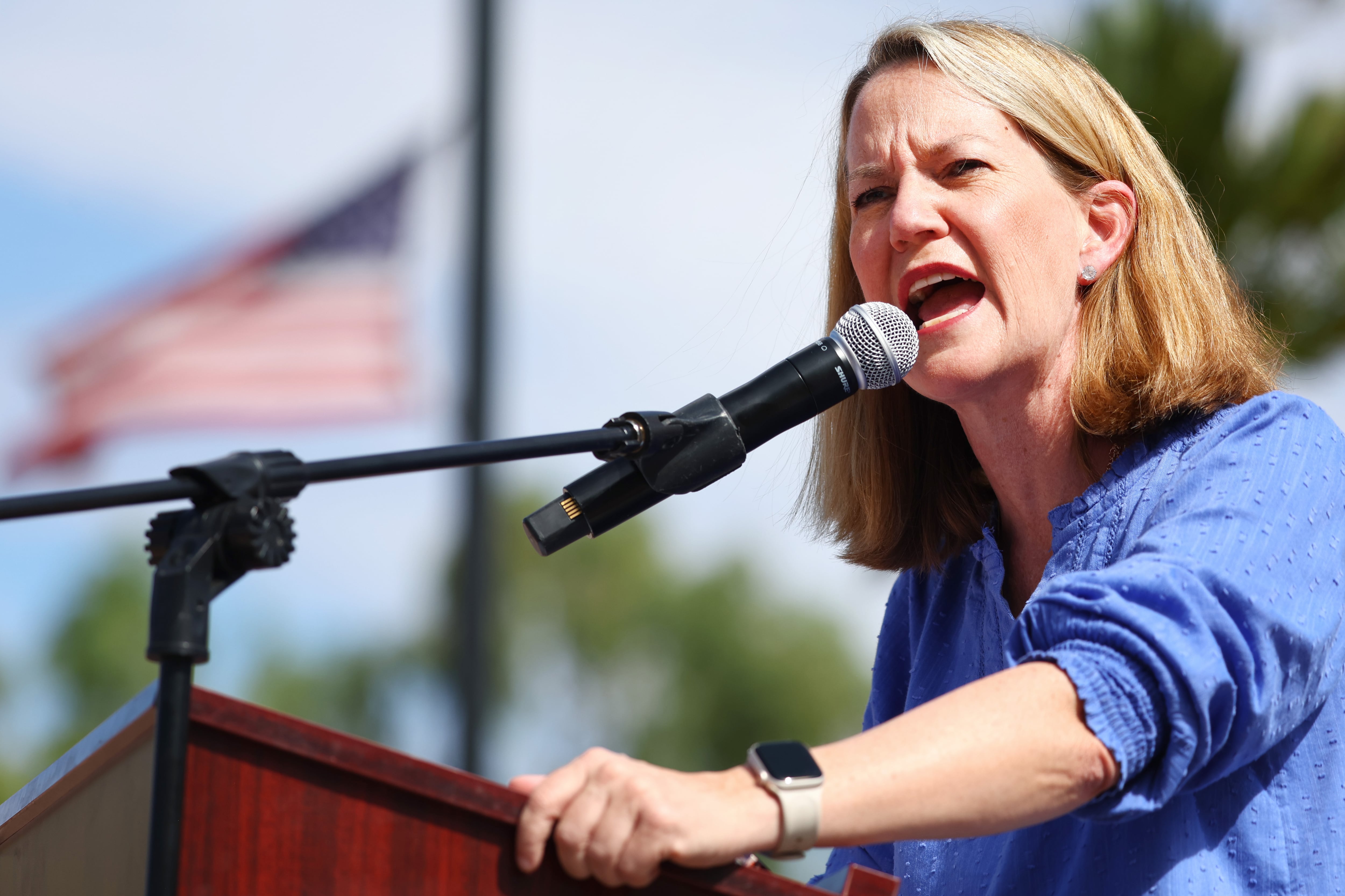 A woman speaks into a microphone outside as an American flag waves in the background