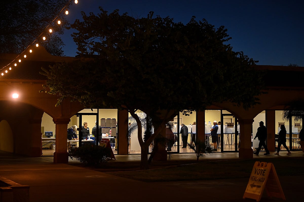 People voting inside of a building with three large windows that are illuminated at night with a string of outdoor lights in the top left corner and a small vote here sign on the bottom right.