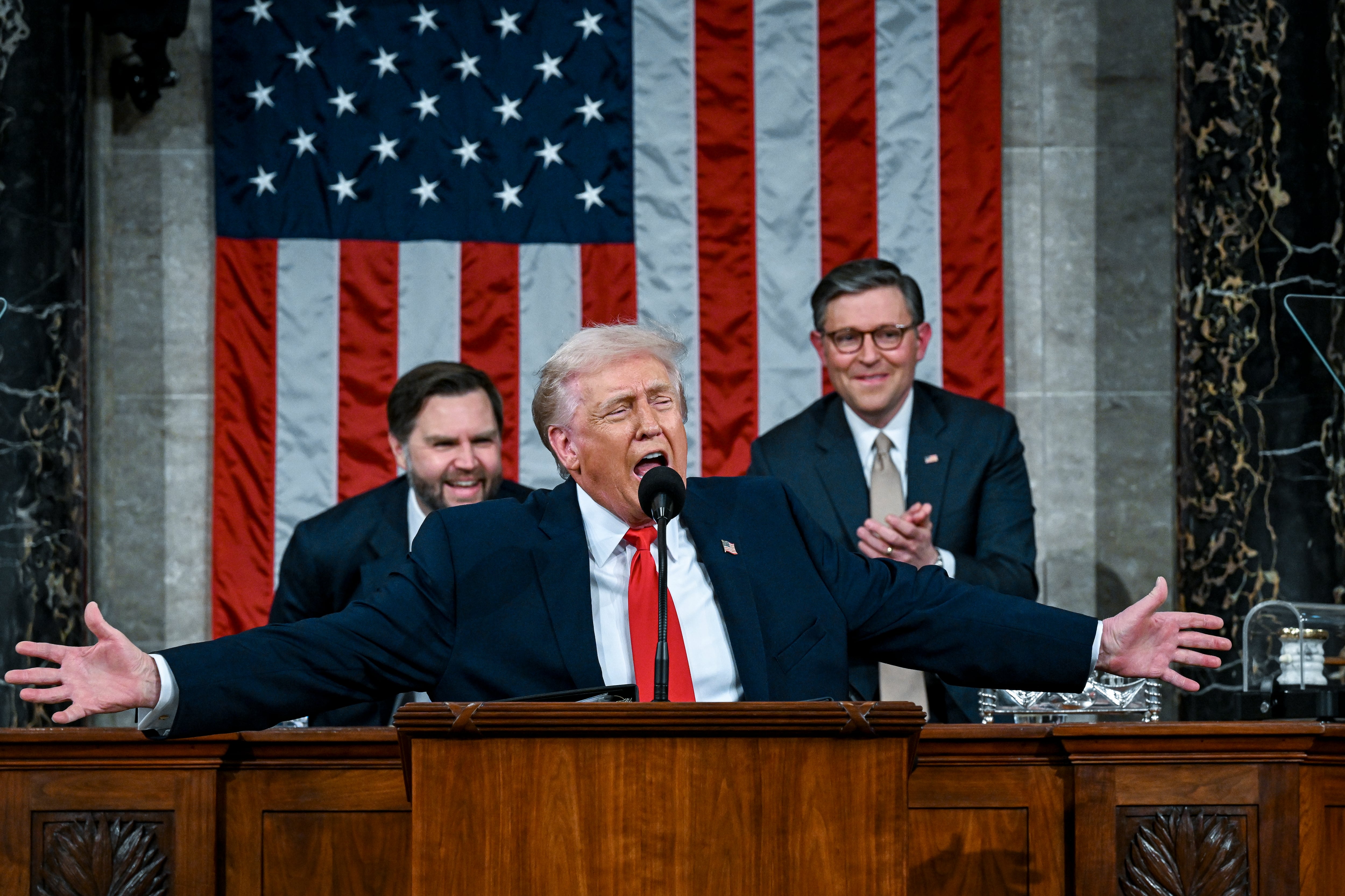 A man in a blue suit jacket and red tie holds his arms out while talking.
